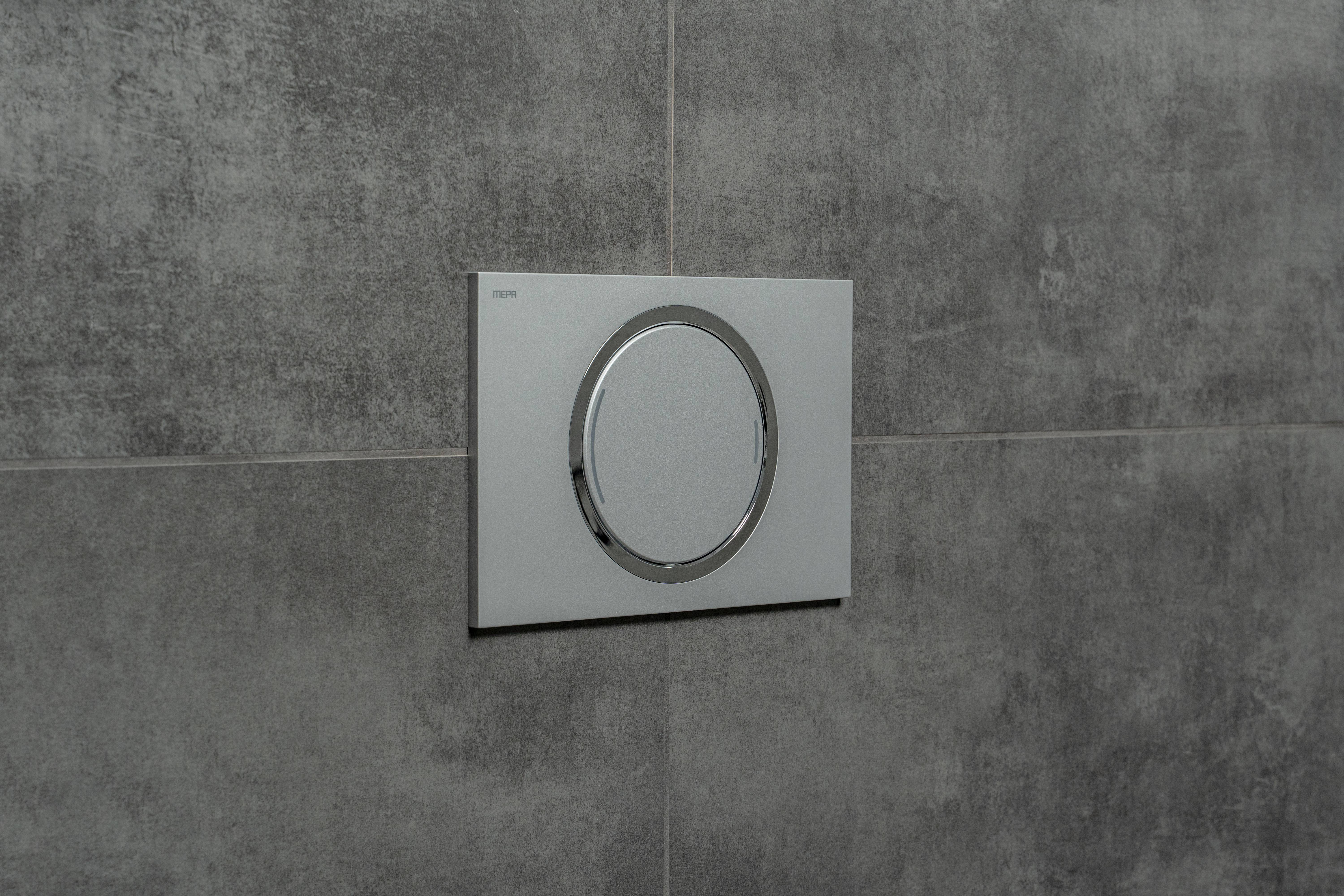 Eine moderne, silberne Betätigungsplatte für die Toilettenspülung ist an einer grauen gefliesten Wand montiert und verfügt über ein kreisförmiges Chrom-Knopfdesign für einen schlichten, minimalistischen Look.