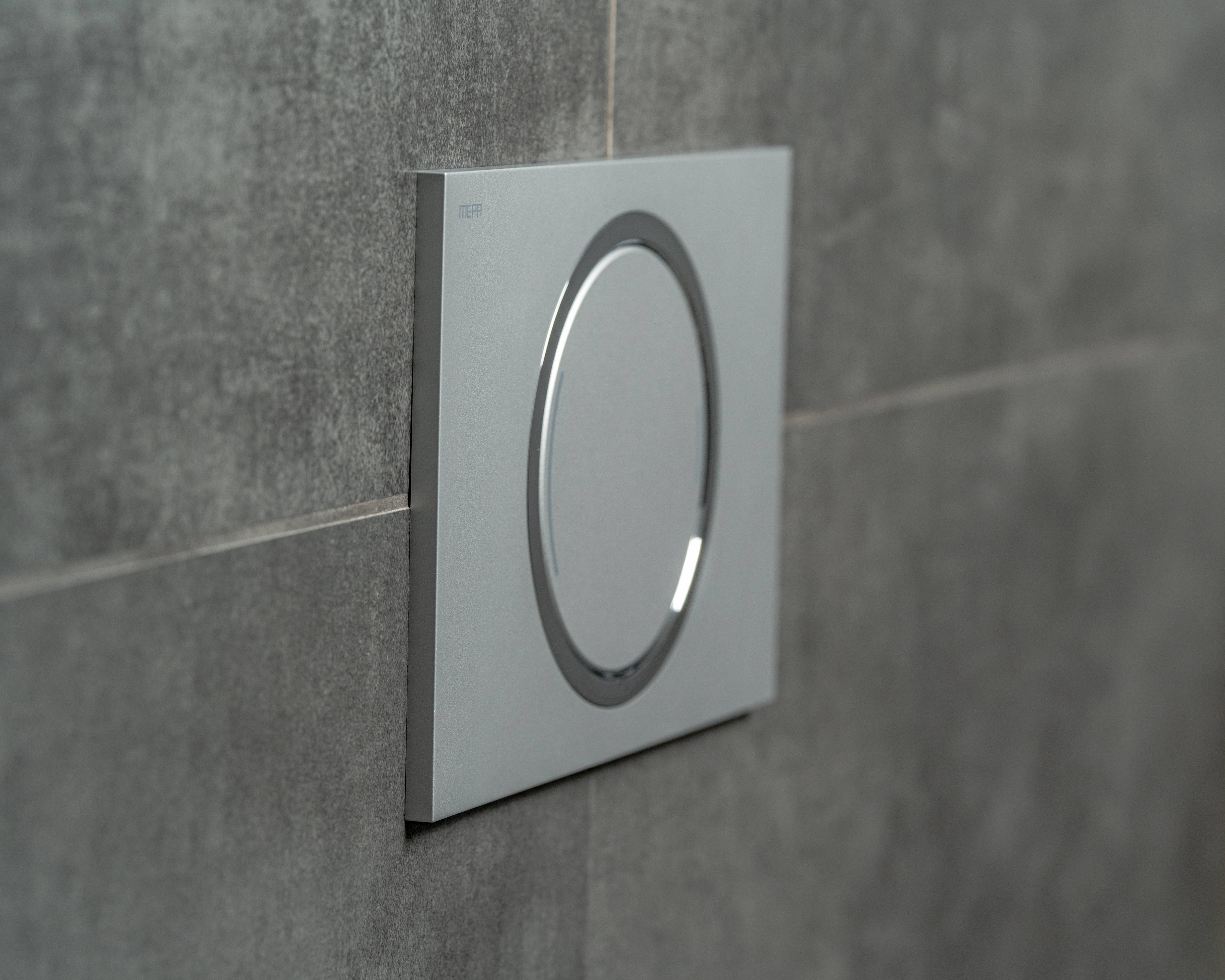 Ein moderner, metallischer Lichtschalter mit kreisförmigem Design ist an einer grauen gefliesten Wand installiert und fügt sich nahtlos in die Dekoration ein.