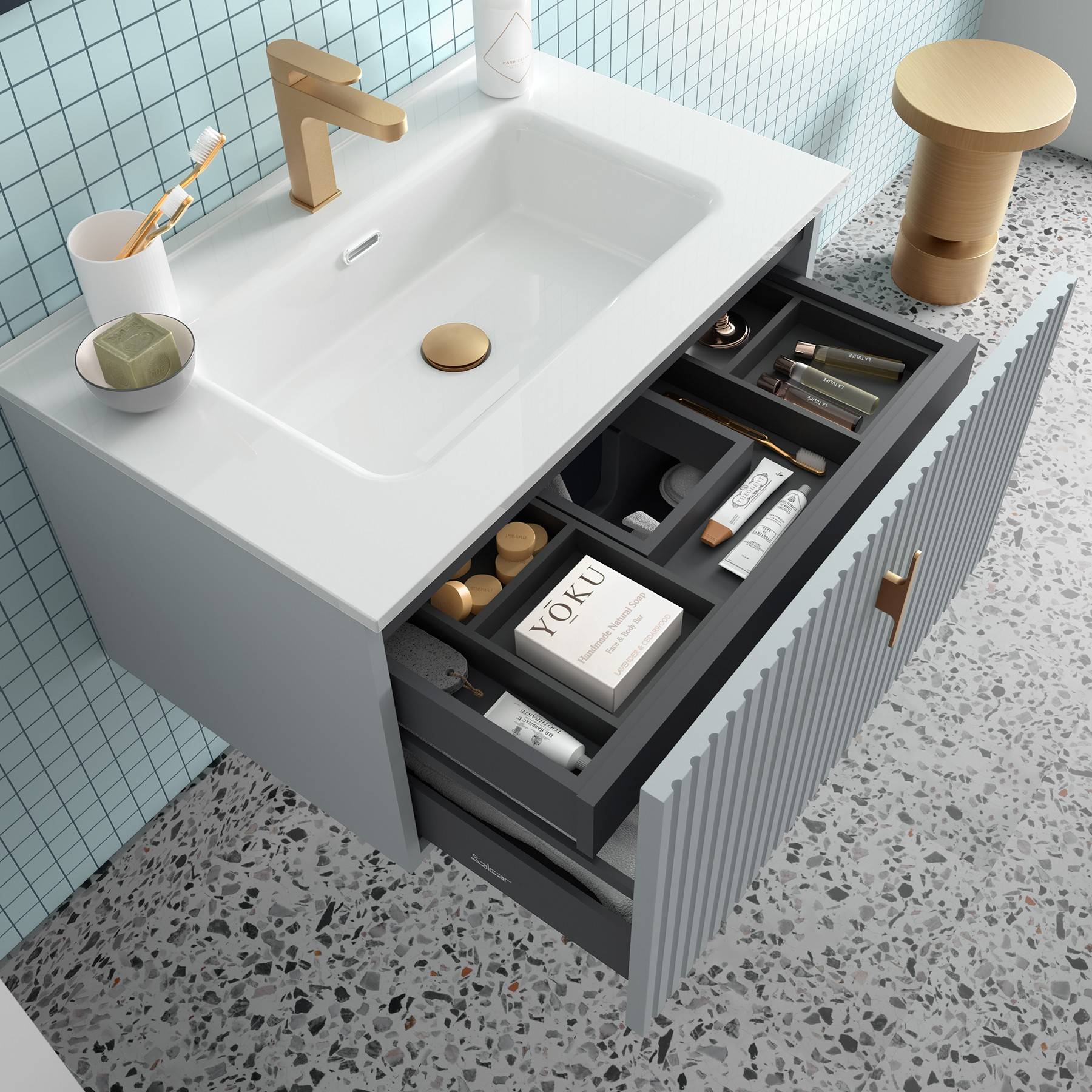 Ein Badezimmer-Waschtisch mit einem weißen Waschbecken, goldenem Wasserhahn und einer geöffneten Schublade, in der Toilettenartikel ordentlich angeordnet sind, vor einem Terrazzoboden und einer gekachelten Wand.