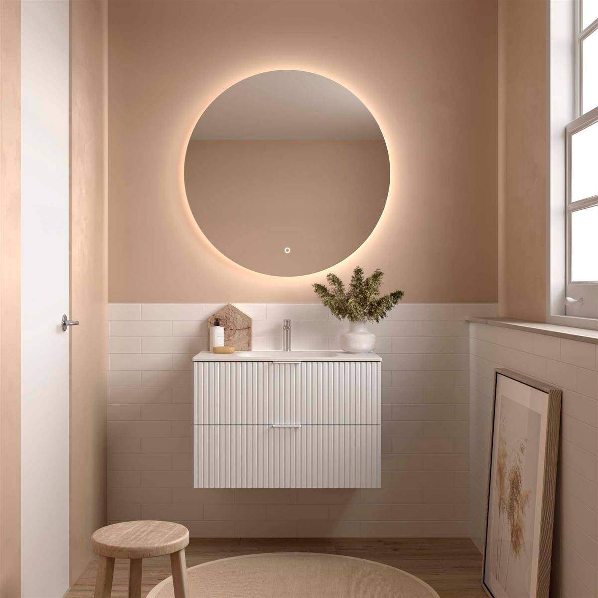 Modernes beiges Badezimmer mit rundem hinterleuchtetem Spiegel über einem weißen Waschtisch, Hocker, Fenster und lehnendem Kunstwerk an der Wand.