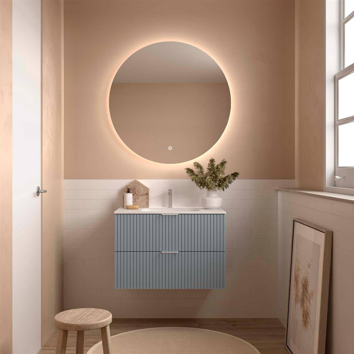 Minimalistes Badezimmer mit rundem hinterleuchtetem Spiegel über blauem Waschtisch, weißen Fliesen, Holzhocker, rahmentem Kunstwerk und Fensterlicht.