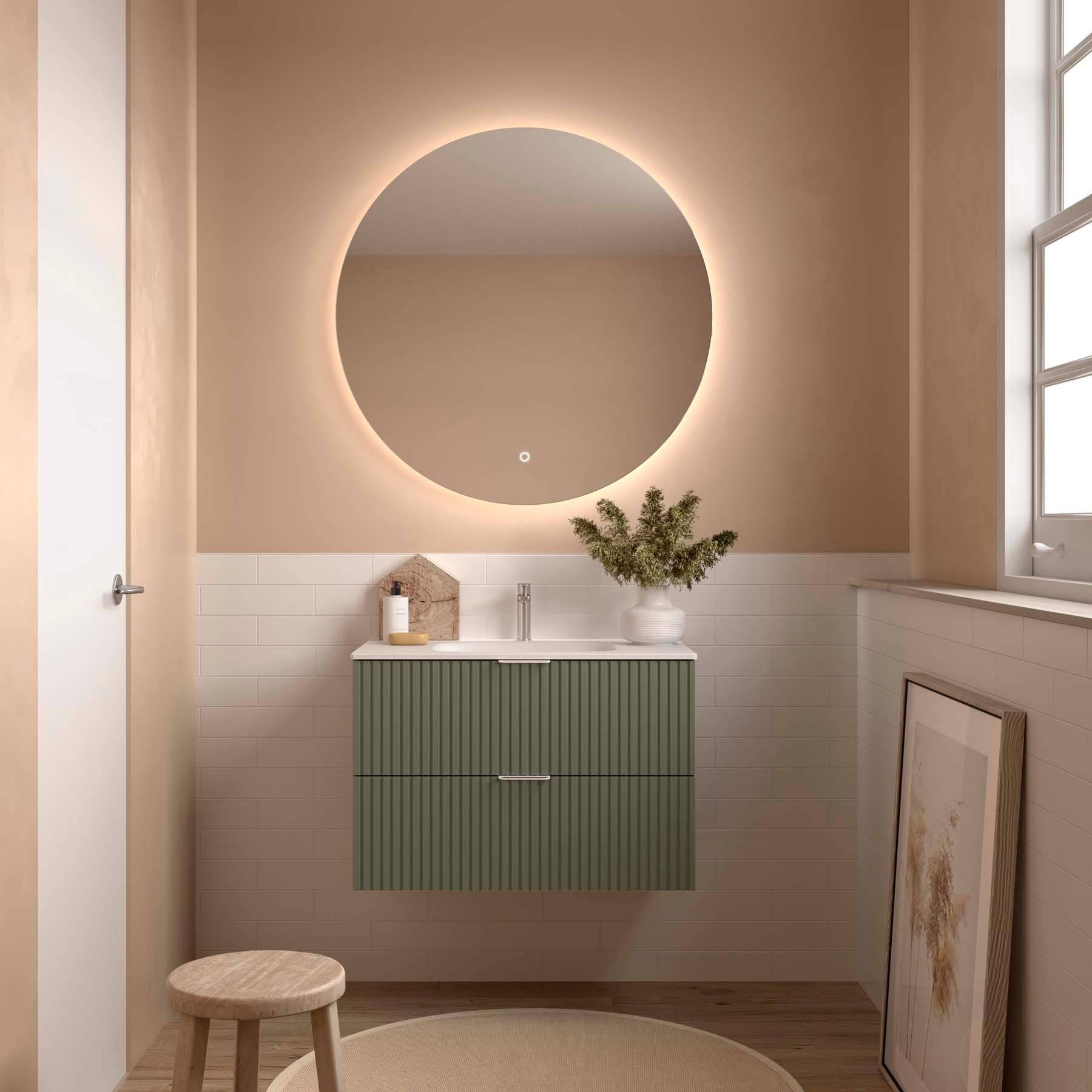 Modernes Badezimmer mit rundem Spiegel und leuchtender Kante. Beige Wände, grüne Unterschrankeinheit unter dem Waschbecken, kleine Pflanze und gerahmte Kunst, die an der Wand lehnt.