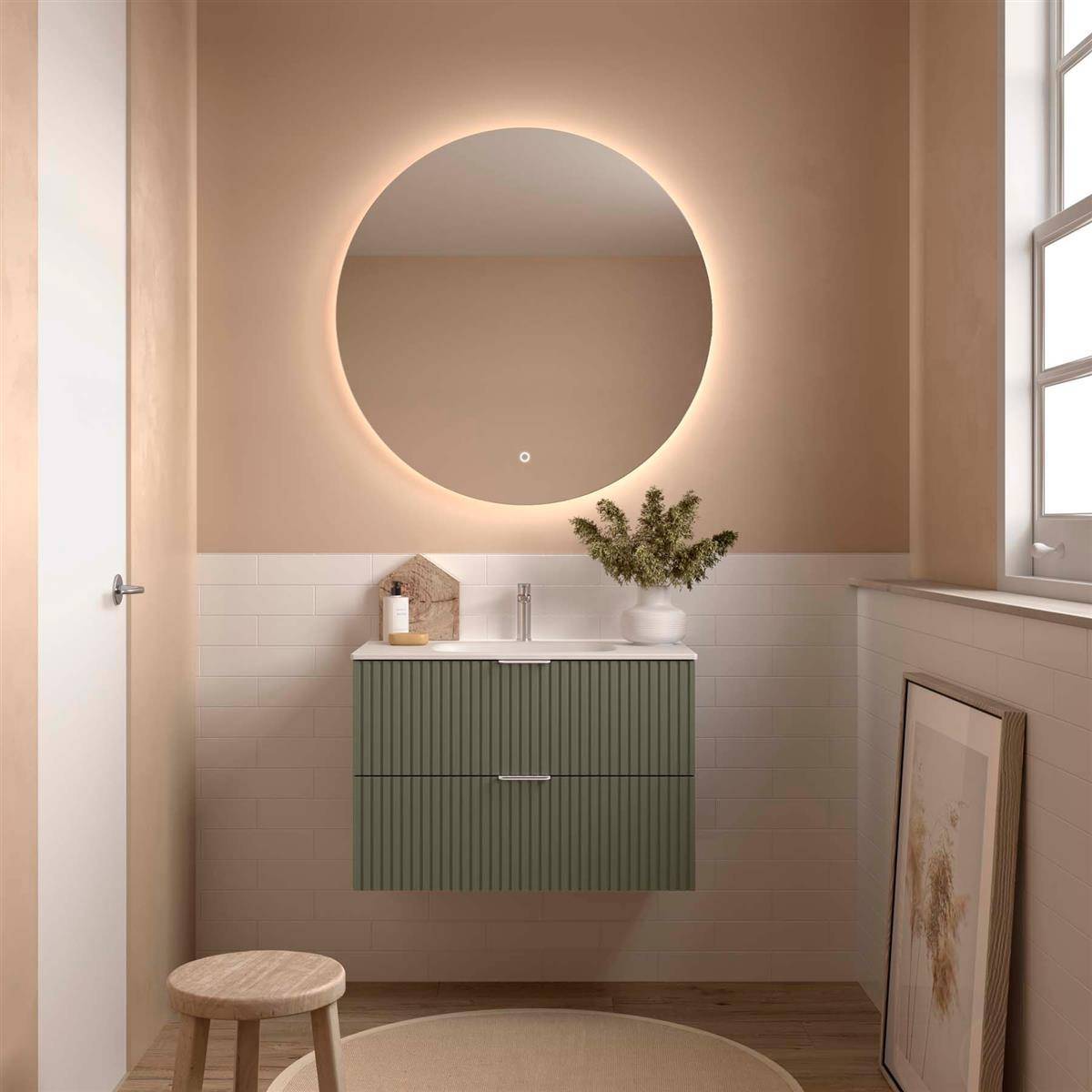 Minimalistisches Badezimmer mit einem runden beleuchteten Spiegel über einem grün wandmontierten Waschtisch, weißem Waschbecken, Holzhocker und angelehntem Kunstwerk am Fenster.