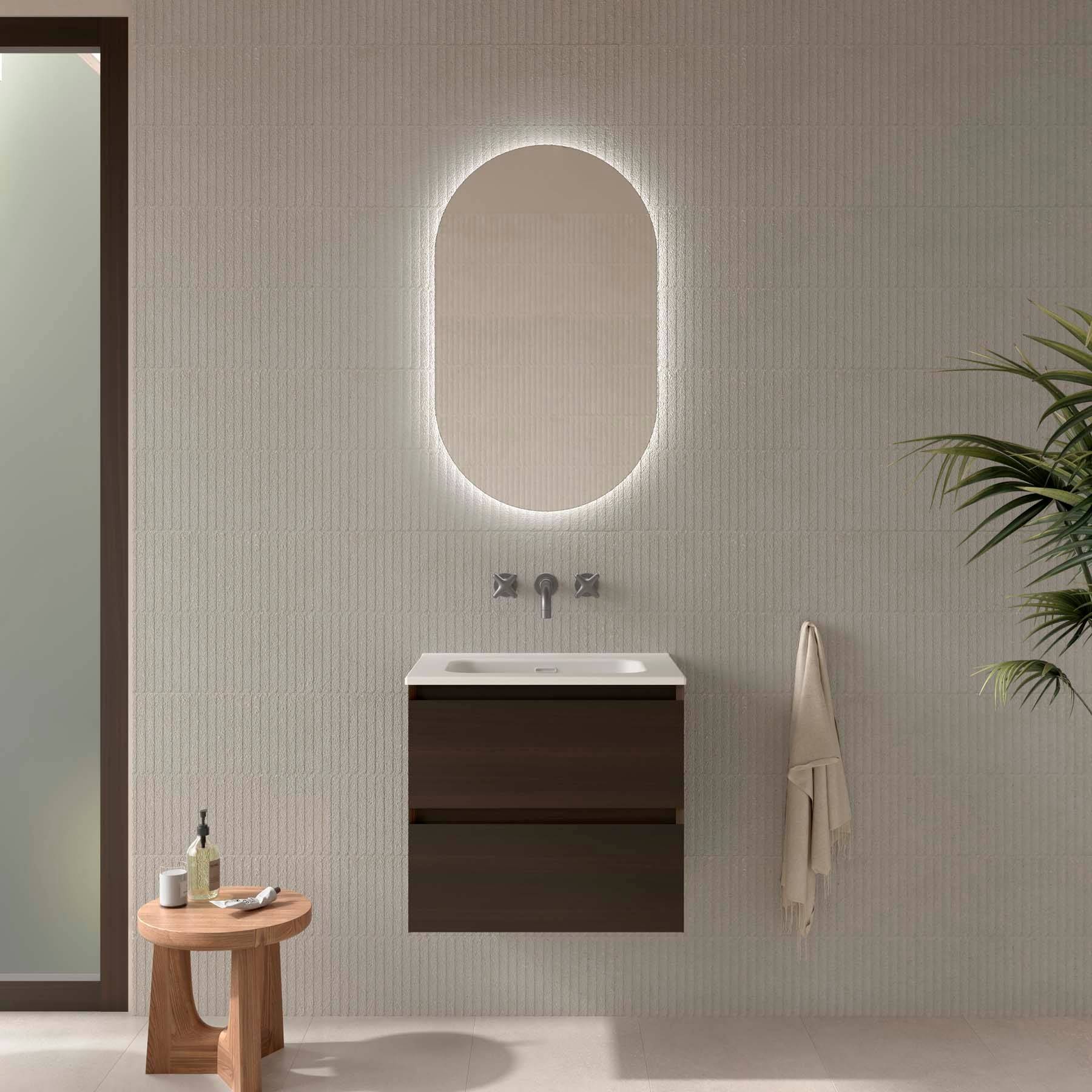 Modernes Badezimmer mit einem ovalen, beleuchteten Spiegel, einem eleganten Waschtisch aus dunklem Holz und einem Handtuch, das rechts an einer hell gefliesten Wand hängt.