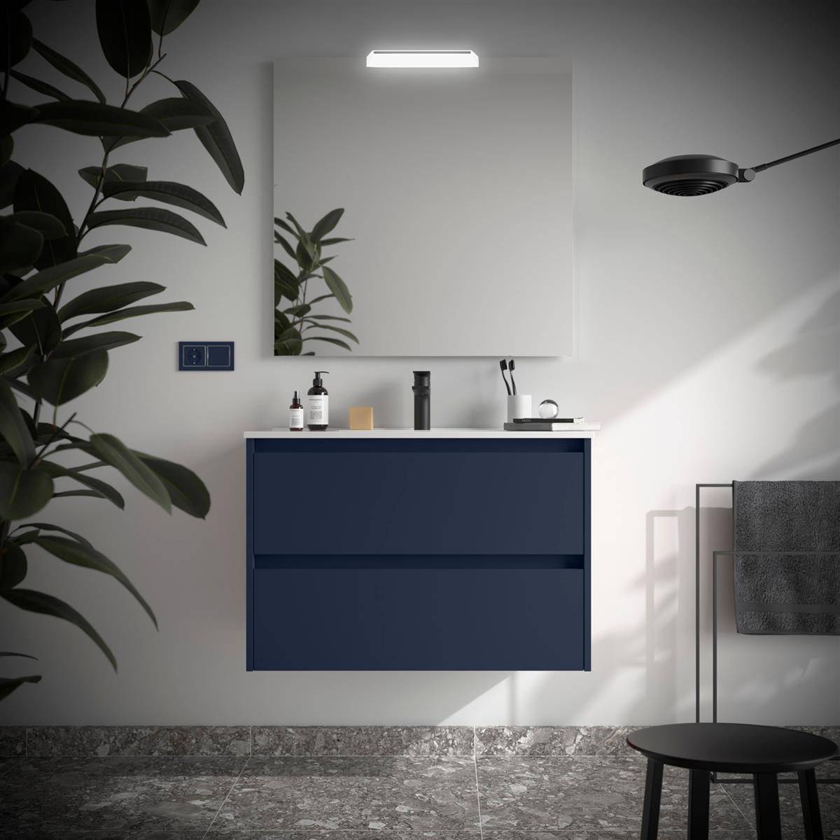 Modernes Badezimmer mit dunkelblauem schwebendem Waschtisch, Spiegel, Wandleuchte, Pflanze, Handtuchhalter und dunklem Hocker auf Steinfliesenboden.