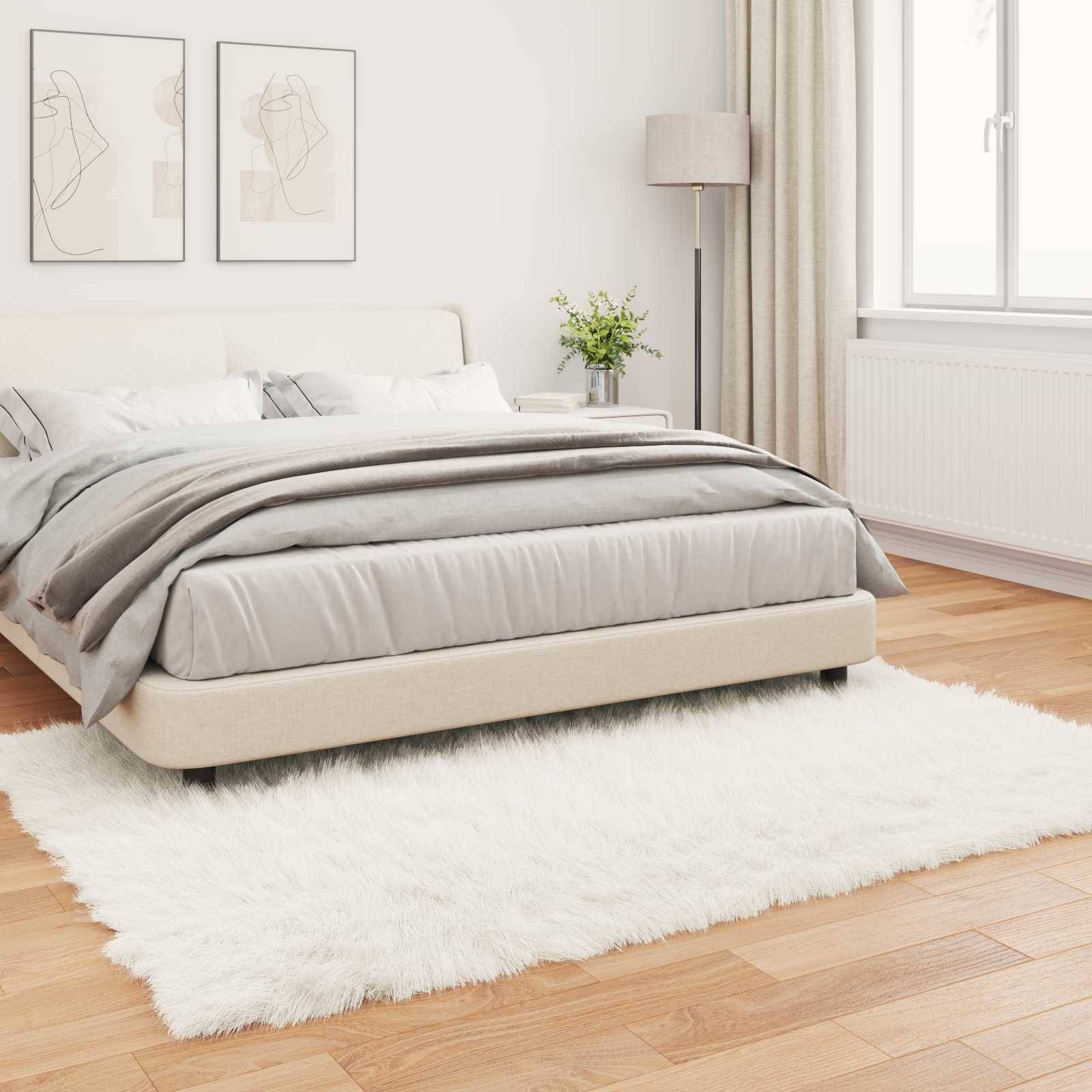 Ein modernes Schlafzimmer mit einem beigen Bett, weißem fluffigem Teppich und hellgrauer Bettwäsche. An der Wand hängen zwei abstrakte Kunstwerke und eine Lampe, und auf dem Nachttisch steht eine Pflanze.