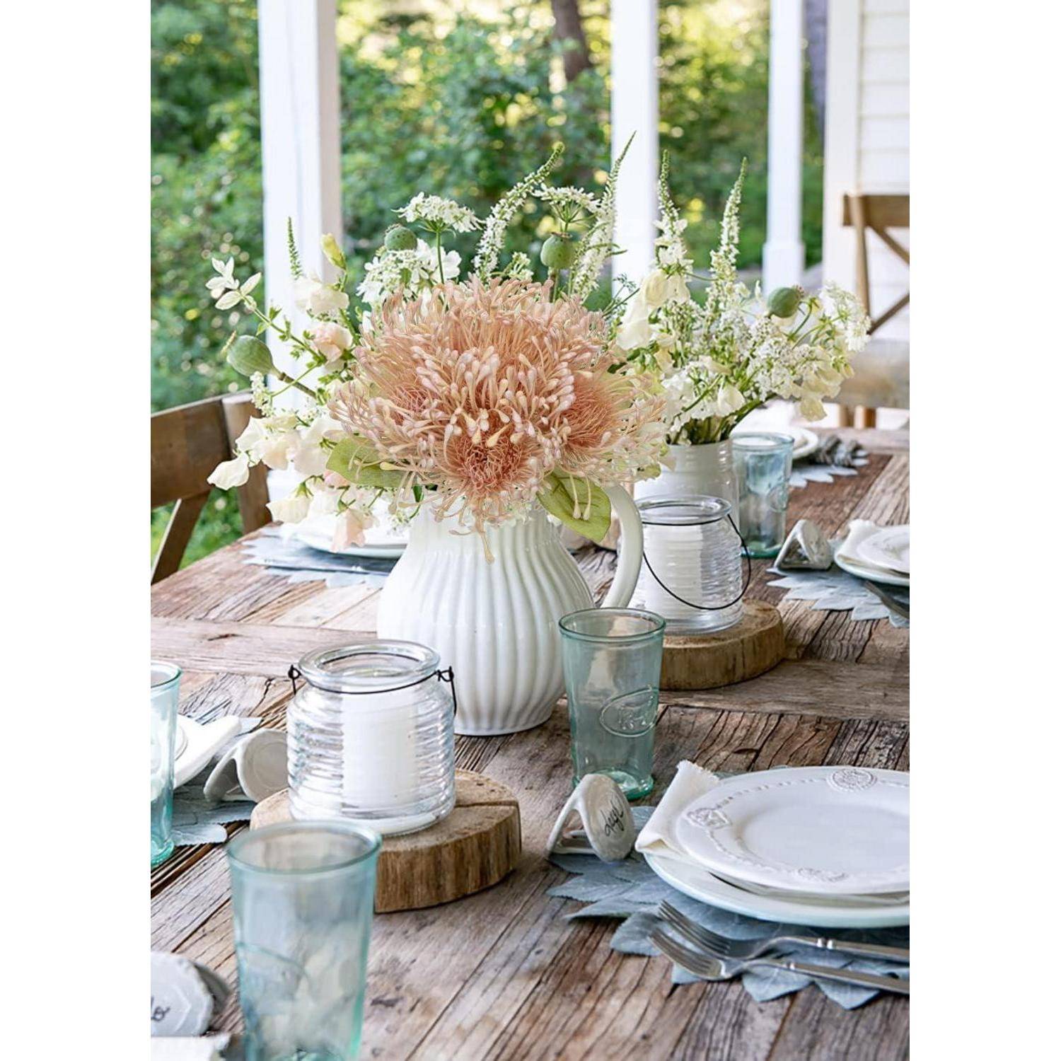 Ein rustikaler Holztisch steht im Freien, gedeckt mit weißen Tellern, blauen Gläsern und einer weißen Vase, die Pfirsich- und weiße Blumen enthält, unter natürlichem Licht.