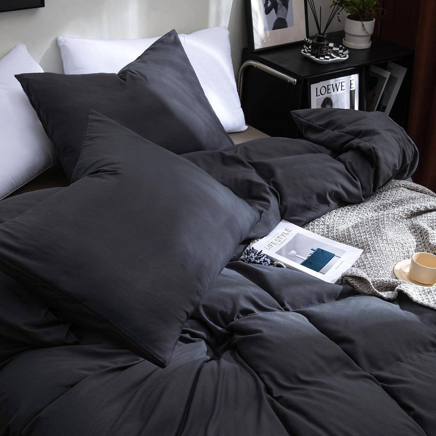 Ein gemütliches, ordentlich hergerichtetes Bett mit dunkelgrauer Bettwäsche, einem weißen Kissen, einer gemusterten Decke, einem Buch und einem hellholzfarbenen Nachttisch in der Nähe.