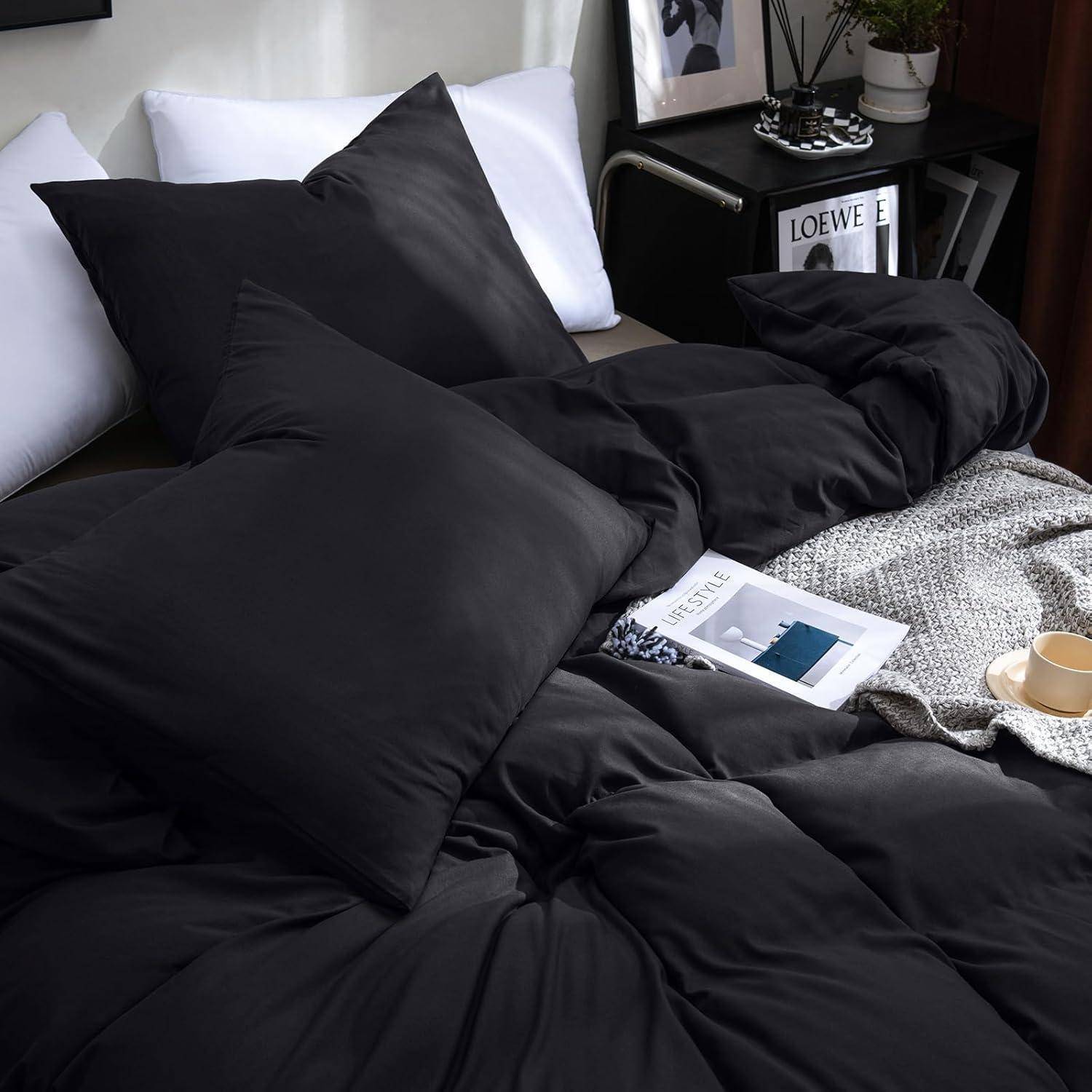 Ein gemütliches Bett mit schwarzen Laken und Kissen, ein gemustertes Buch und eine graue gestrickte Decke. Ein Schreibtisch mit Büchern und einem gerahmten Foto im Hintergrund.