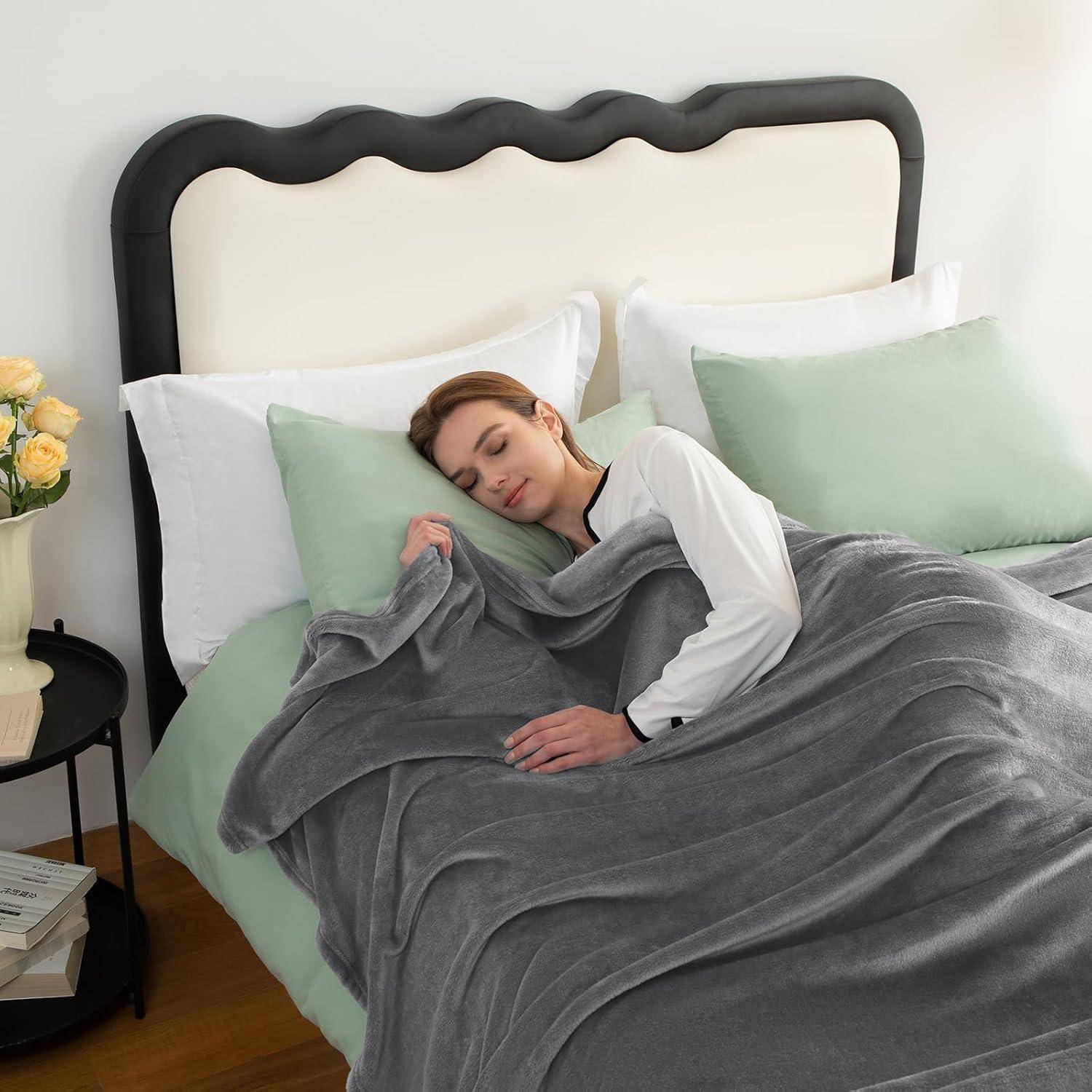 Eine Person, die friedlich auf einem Bett mit weiß-grüner Bettwäsche schläft, bedeckt von einer grauen Decke, neben einem Nachttisch mit einer Pflanze und Büchern.