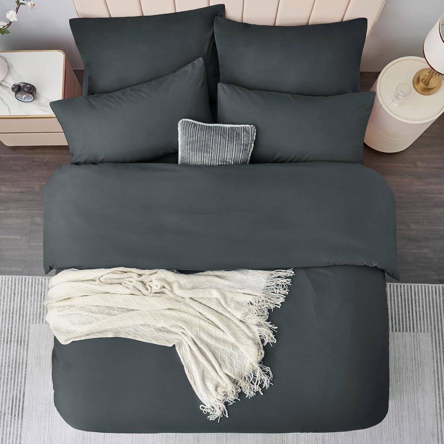 Ein ordentlich gemachtes Bett mit dunkelgrauer Bettwäsche, einem kleinen grauen gestreiften Kissen und einer beige farbenen Decke. Daneben stehen ein Nachttisch und eine Lampe.