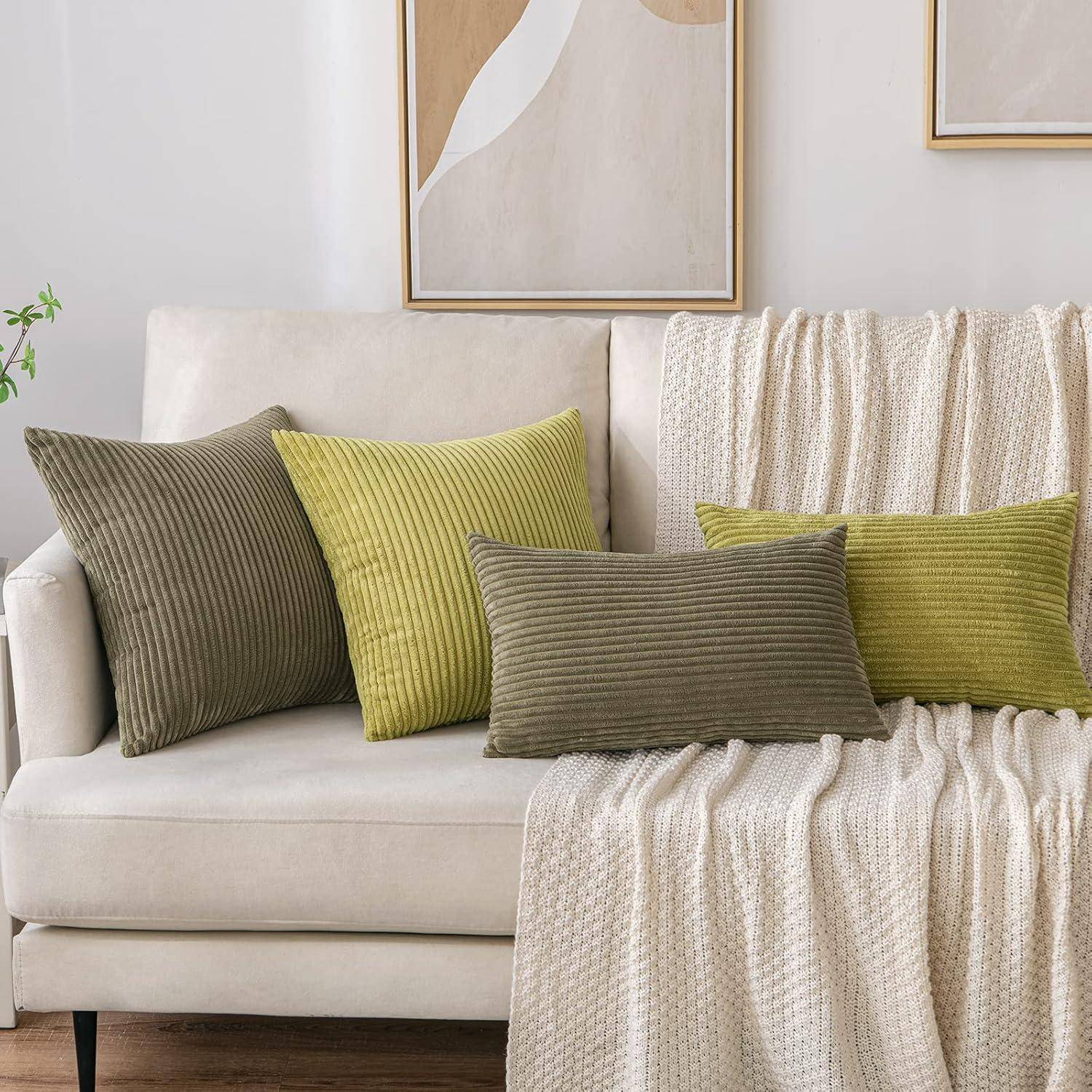 Eine gemütliche cremefarbene Couch ist mit gerippten Zierkissen in Oliv- und Limonengrün ausgestattet. Eine passende strukturierte beige Decke liegt über der Rückenlehne gebreitet. Neutrale Wandkunst ist im Hintergrund sichtbar.