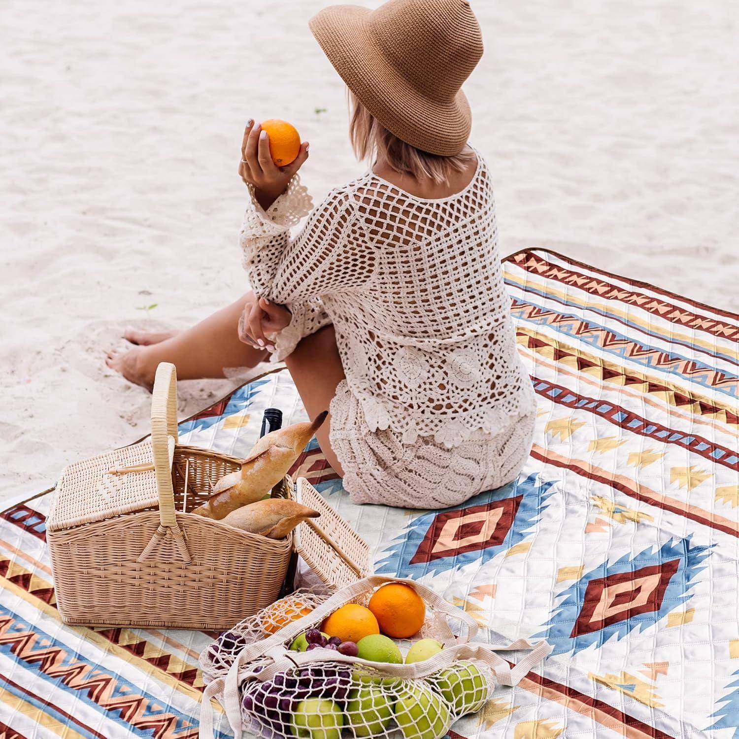 Eine Person sitzt auf einer bunten Strandmatte, trägt einen Strohhut und einen gehäkelten Überwurf und hält eine Orange. Ein Picknickkorb und Früchte befinden sich in der Nähe.