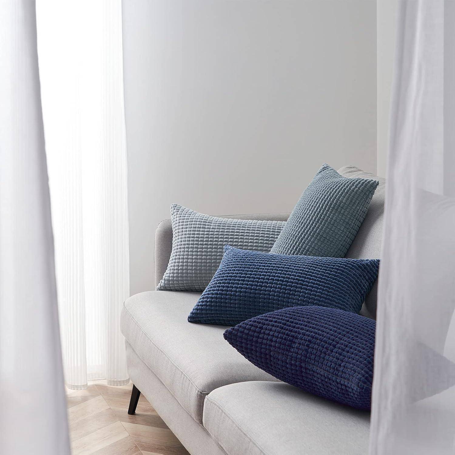 Ein hellgrauer Sofa mit drei strukturierten Kissen in Grau, Blau und Tiefblau steht in der Nähe eines durchsichtigen Vorhangs, durch den weiches Tageslicht hindurchfiltert.