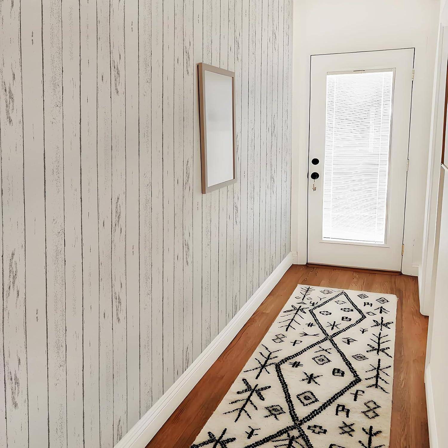 Ein schmaler, lichtdurchfluteter Flur mit Holzböden zeigt einen geometrisch gemusterten Teppich und einen gerahmten Spiegel an der linken Wand, der zu einer Glastür führt.