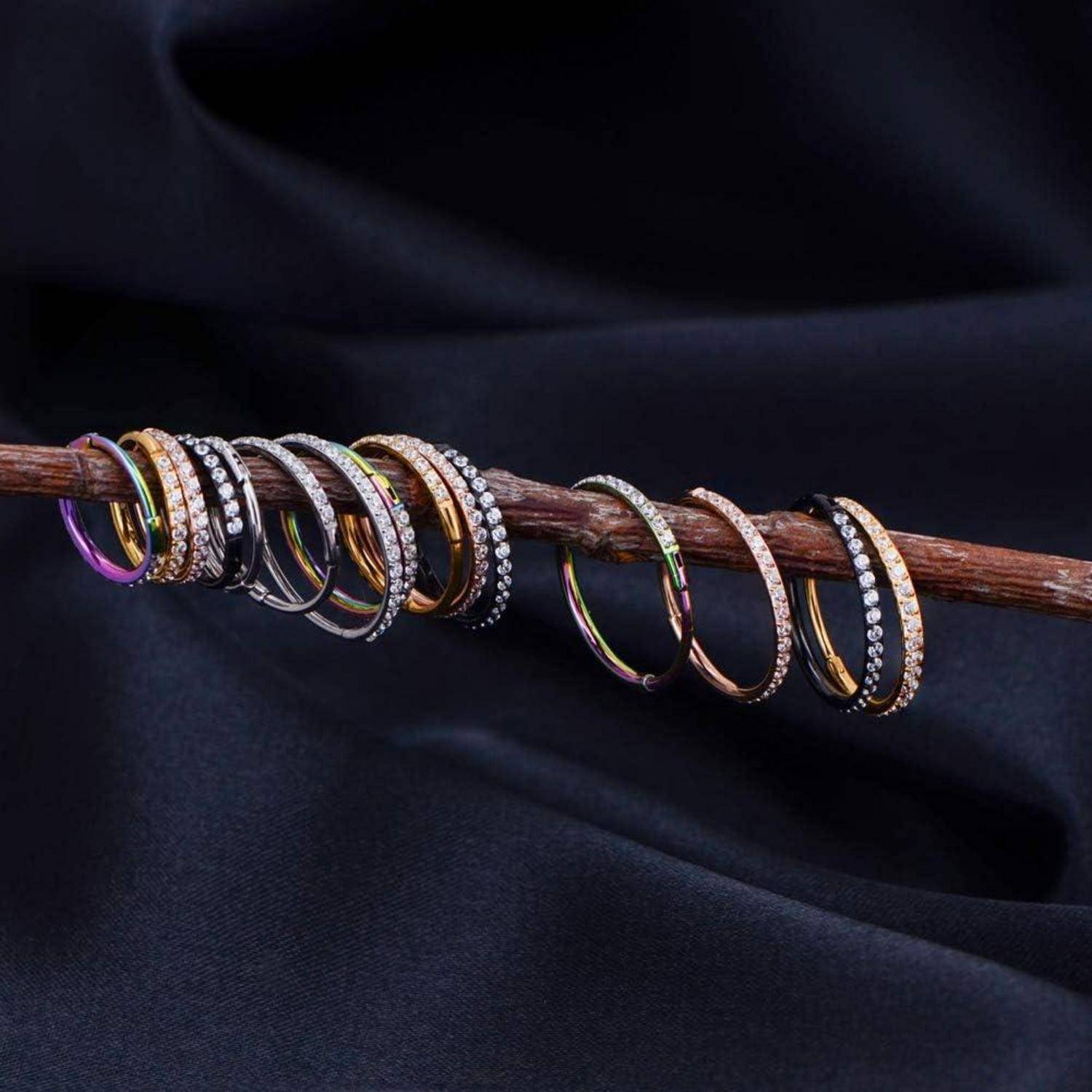 Eine Sammlung glänzender Ringe, einige mit eingesetzten Steinen, nebeneinander auf einer Holzstange vor einem dunklen Stoffhintergrund angeordnet.