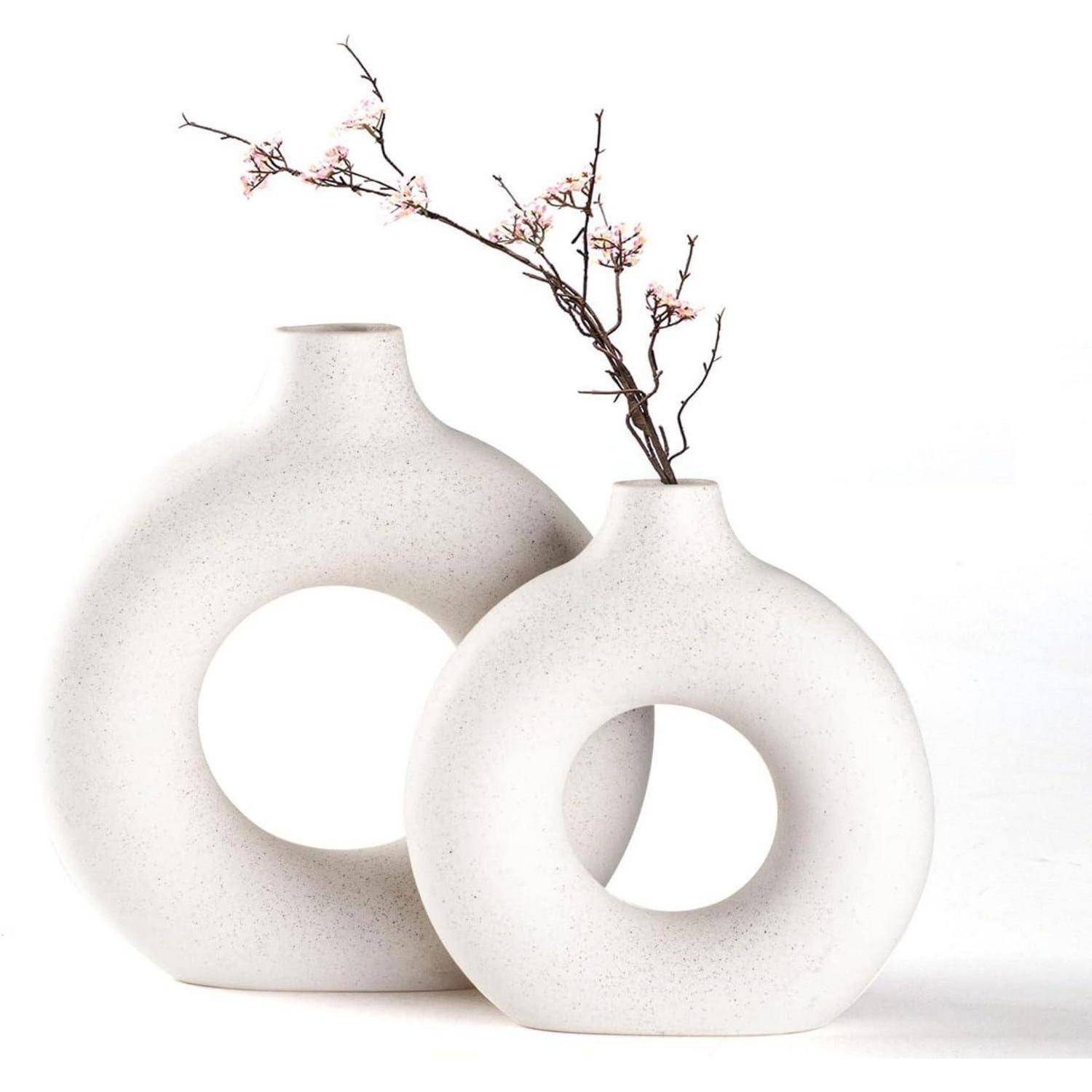 Zwei runde, weiße Keramikvasen mit zentralen Öffnungen; eine davon enthält einen Zweig mit kleinen rosa Blüten. Minimalistisches Design.
