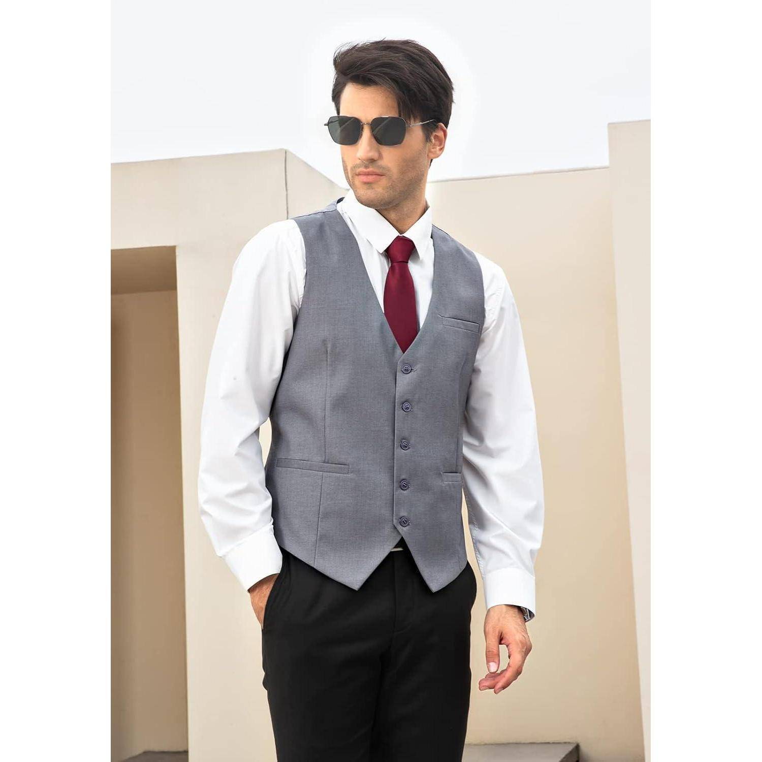 Eine Person mit Sonnenbrille, trägt eine graue Weste über einem weißen Hemd und eine rote Krawatte, steht vor einem modernen beige-weißen architektonischen Hintergrund.