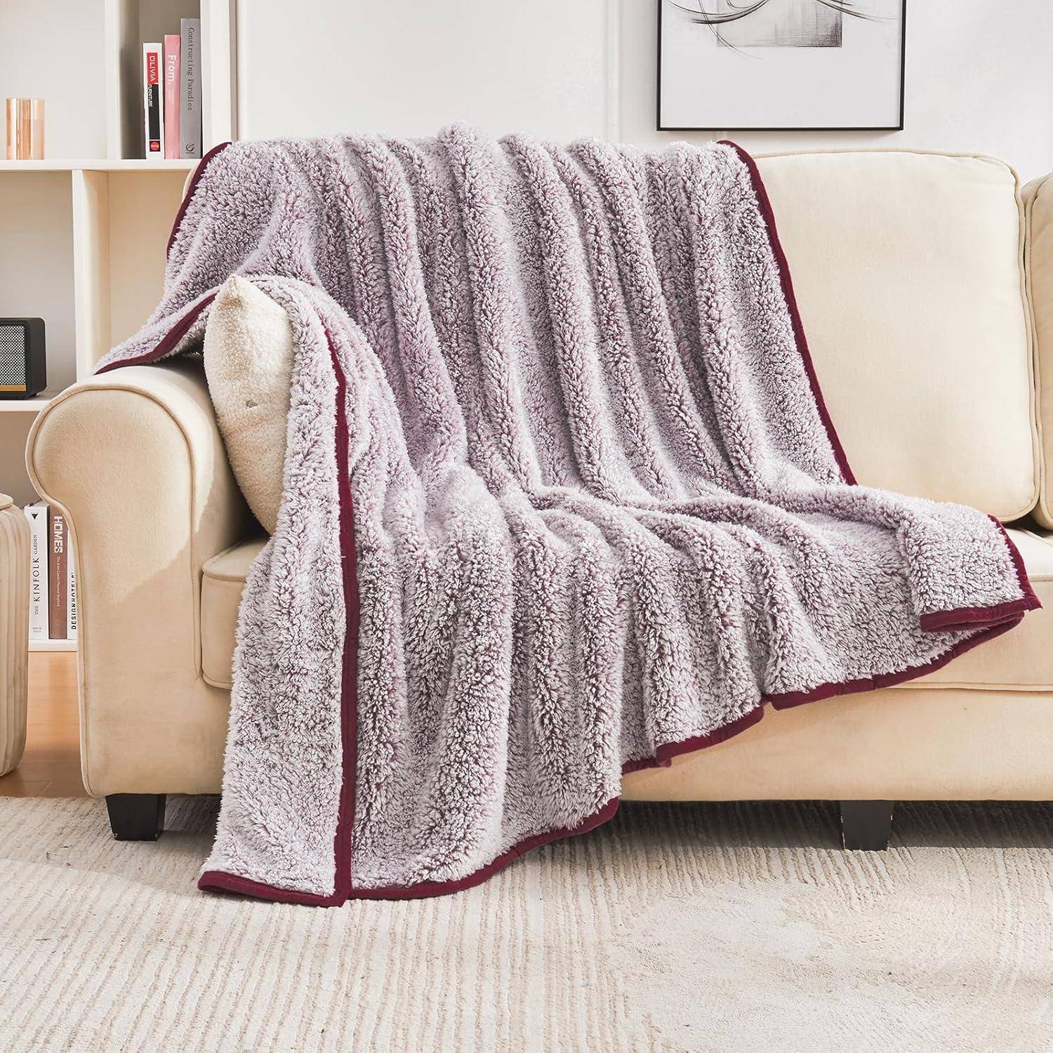Eine weiche, graue Fleece-Decke mit marronroten Kanten, die über einem beigen Sofa in einer gemütlichen Wohnzimmereinrichtung neben einem Bücherregal drapiert liegt.