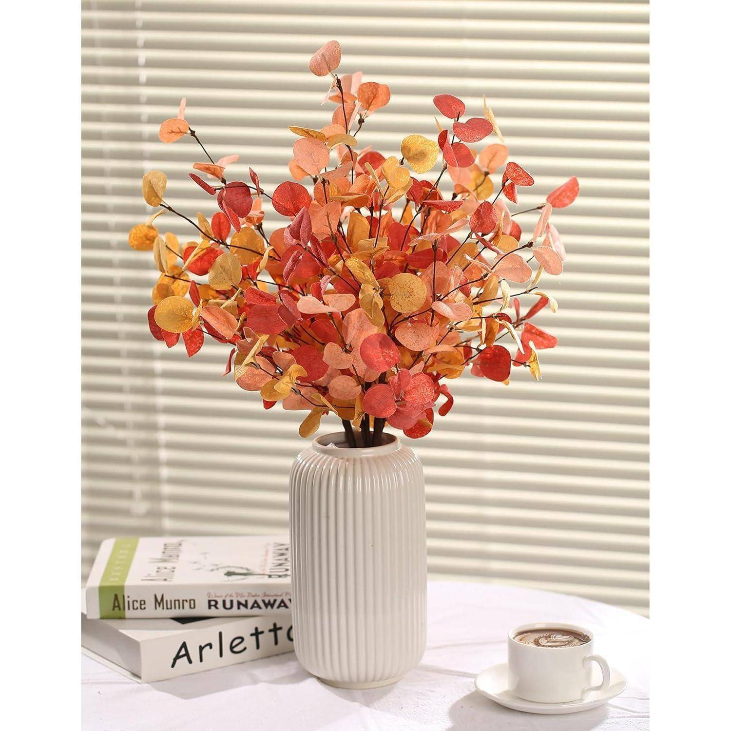 Eine weiße Vase enthält leuchtend orange und gelbe Blätter auf einem Tisch mit Büchern von Alice Munro und Arletto, neben einer Tasse Kaffee.