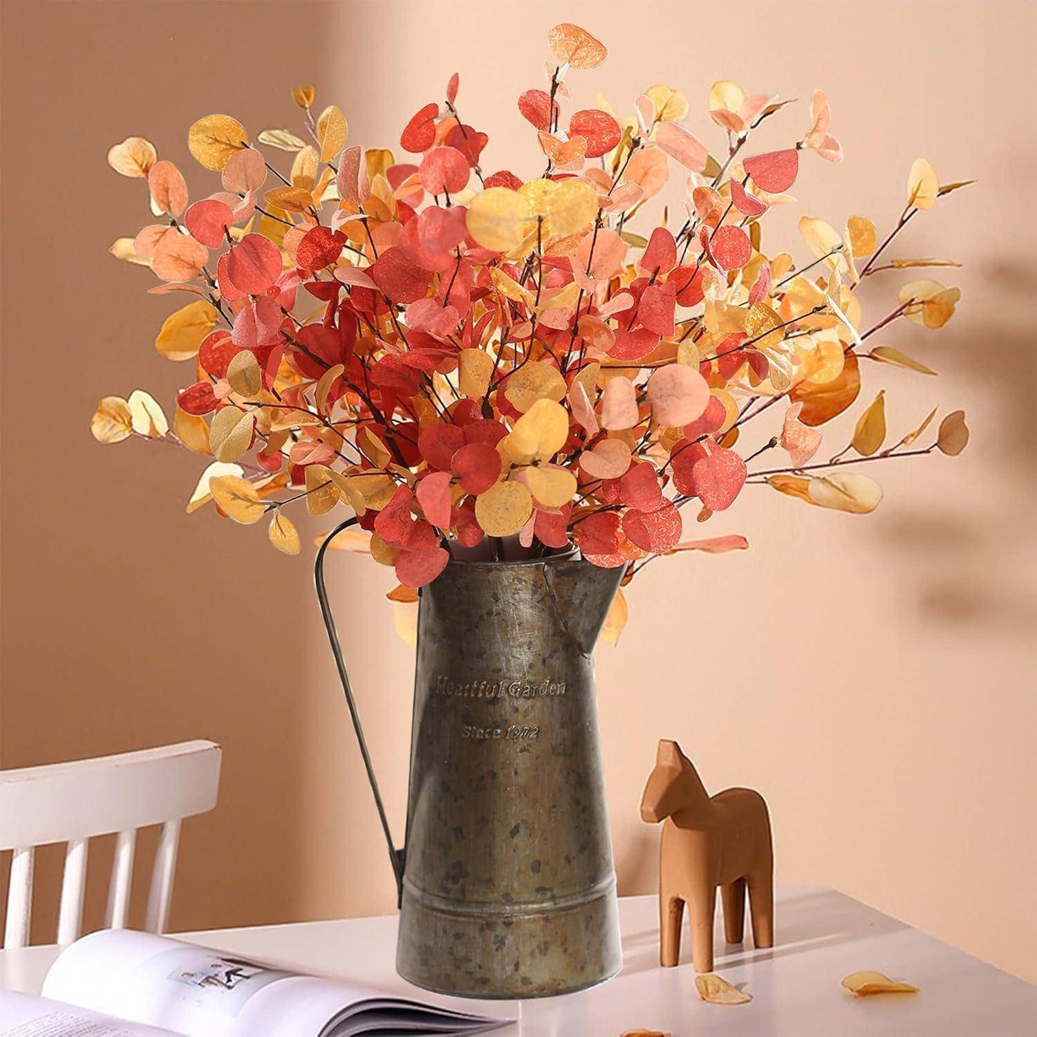 Herbstfarben-gefärbte Blätter, die in einem hohen, rustikalen Metallkrug auf einem Tisch angeordnet sind, mit einem kleinen Holzpferd und einem Stuhl in der Nähe. Warme Töne.
