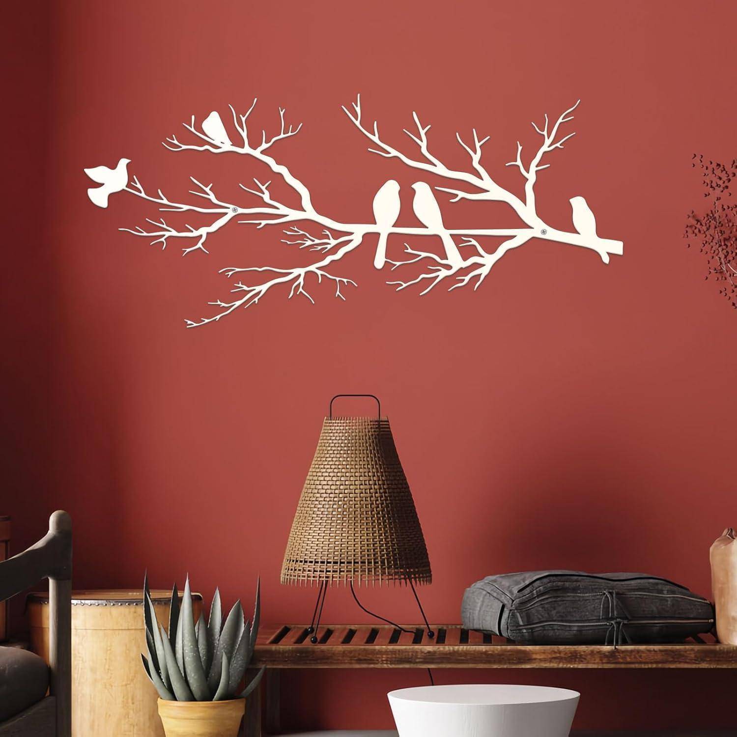 Ein weißes Wandtattoo mit Vögeln auf einem Ast schmückt eine rote Wand über einem gewebten Lampenschirm, daneben eine Kaktuspflanze und ein grauer Kissen.
