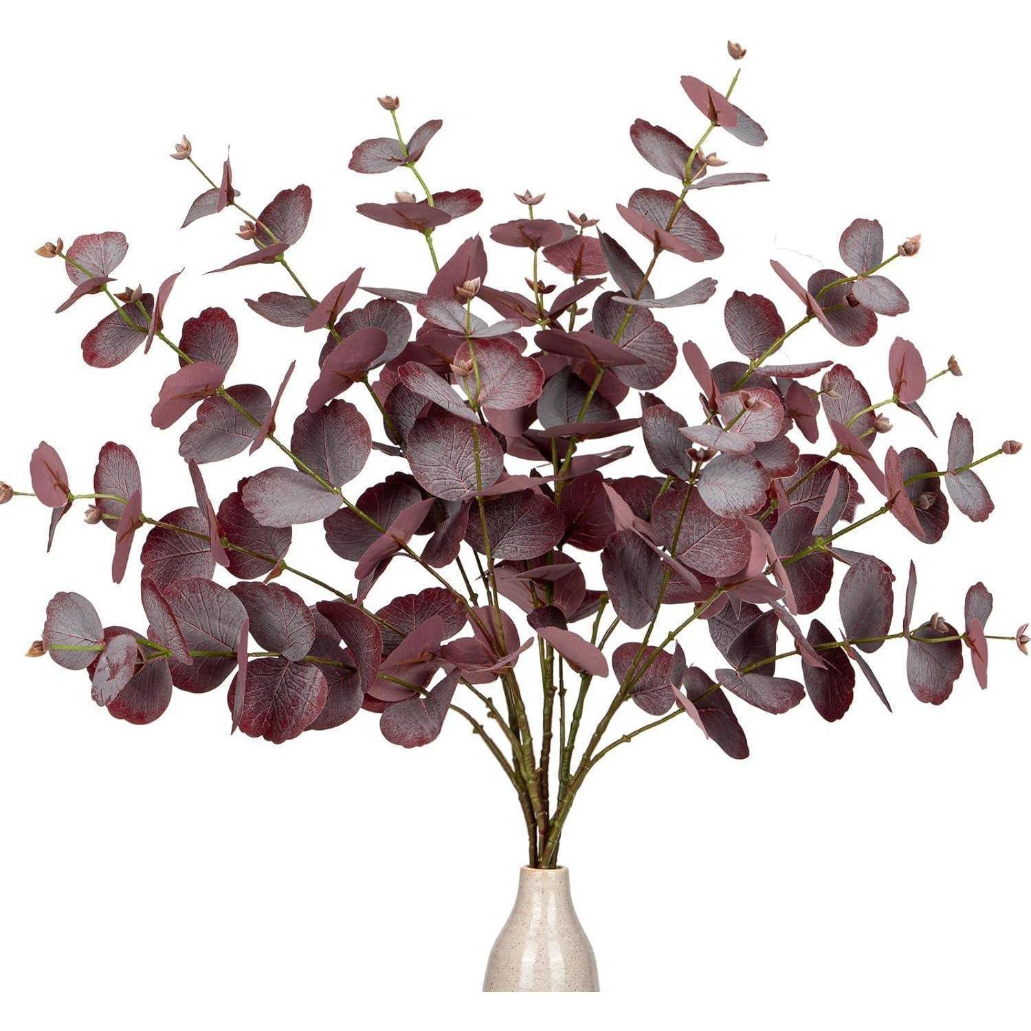Ein Strauß getrockneter Eukalyptuszweige mit dunkelburgundeyfarbenen Blättern, der in einer schlichten beigen Vase präsentiert wird.