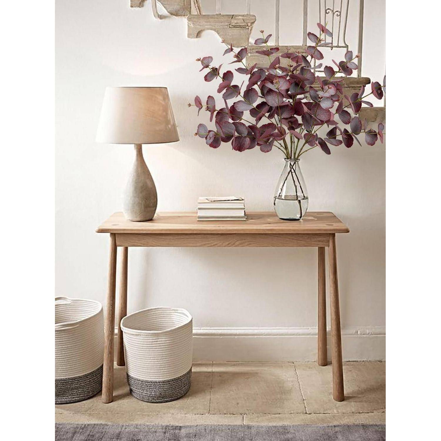Ein Konsolentisch aus Holz mit einer Lampe, Büchern und einer Vase mit lila Blättern. Zwei geflochtene Körbe stehen darunter, und im Hintergrund befinden sich Treppenstufen.