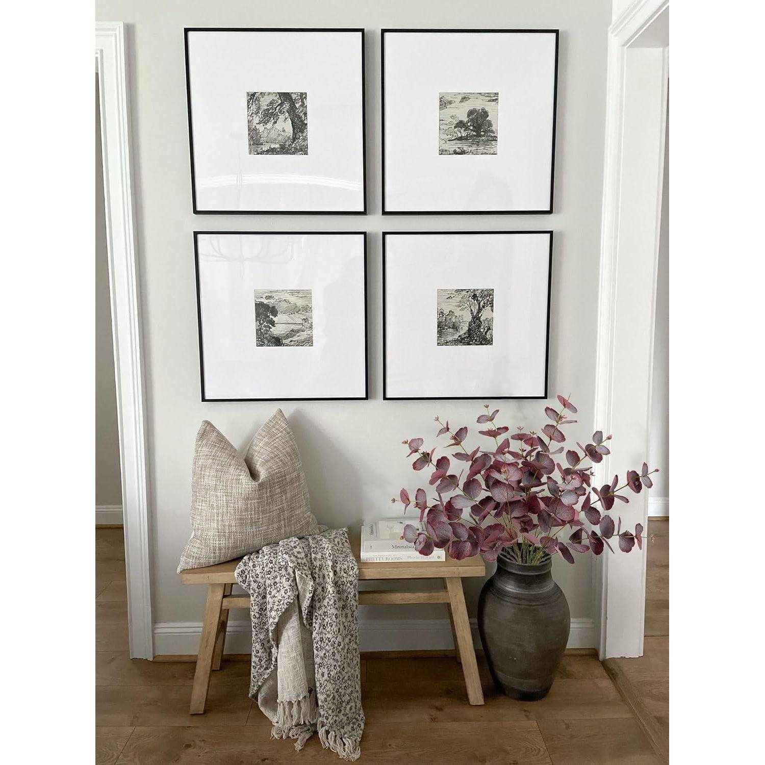 Eine Holzbank mit einem beigen Kissen und einer gemusterten Decke steht unterhalb von vier gerahmten Kunstwerken an der Wand. Eine Vase mit rosa Blattwerk befindet sich neben der Bank.