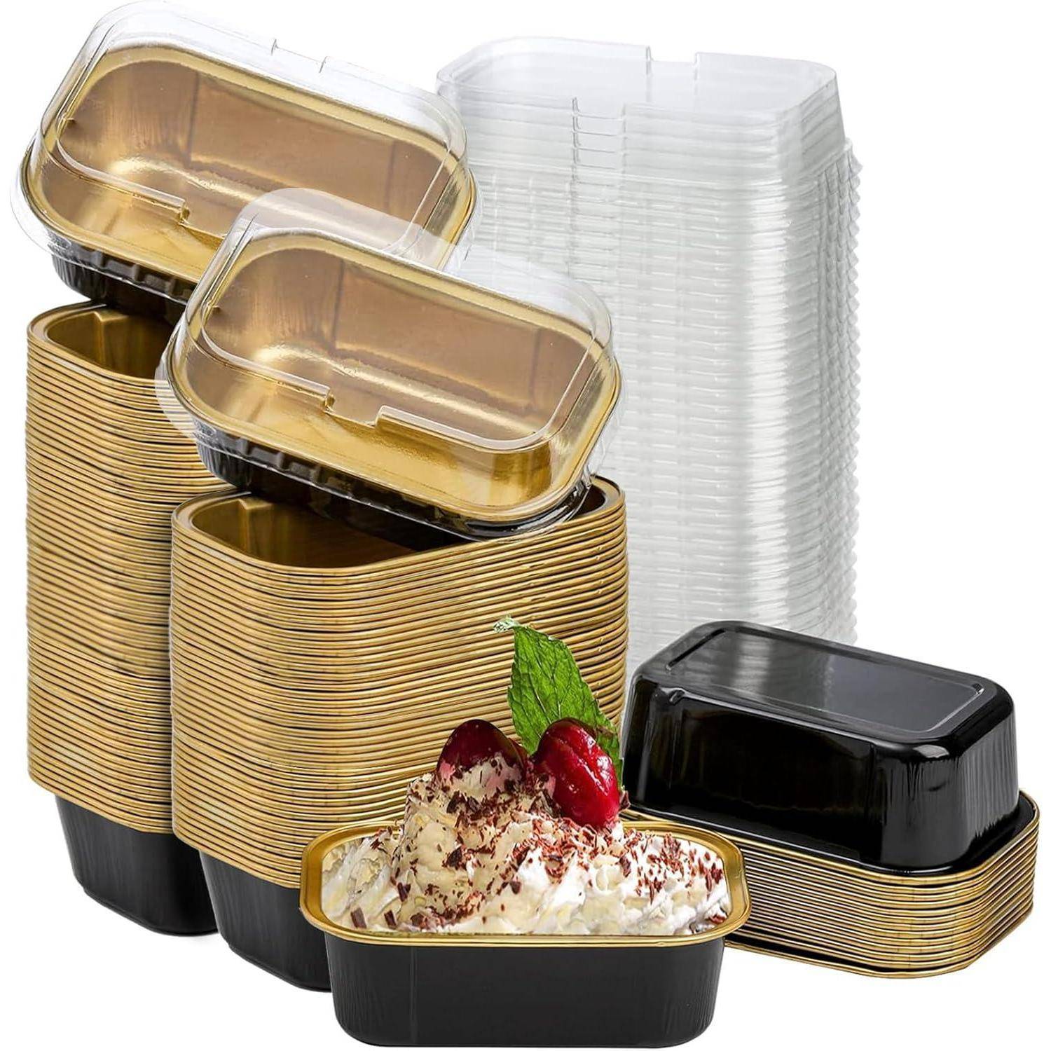 Ein Stapel von Gold- und schwarzen Lebensmittelbehältern mit Deckel neben einem Dessert, das mit Schokoladenspänen und Kirschen auf einem der Behälter verziert ist.