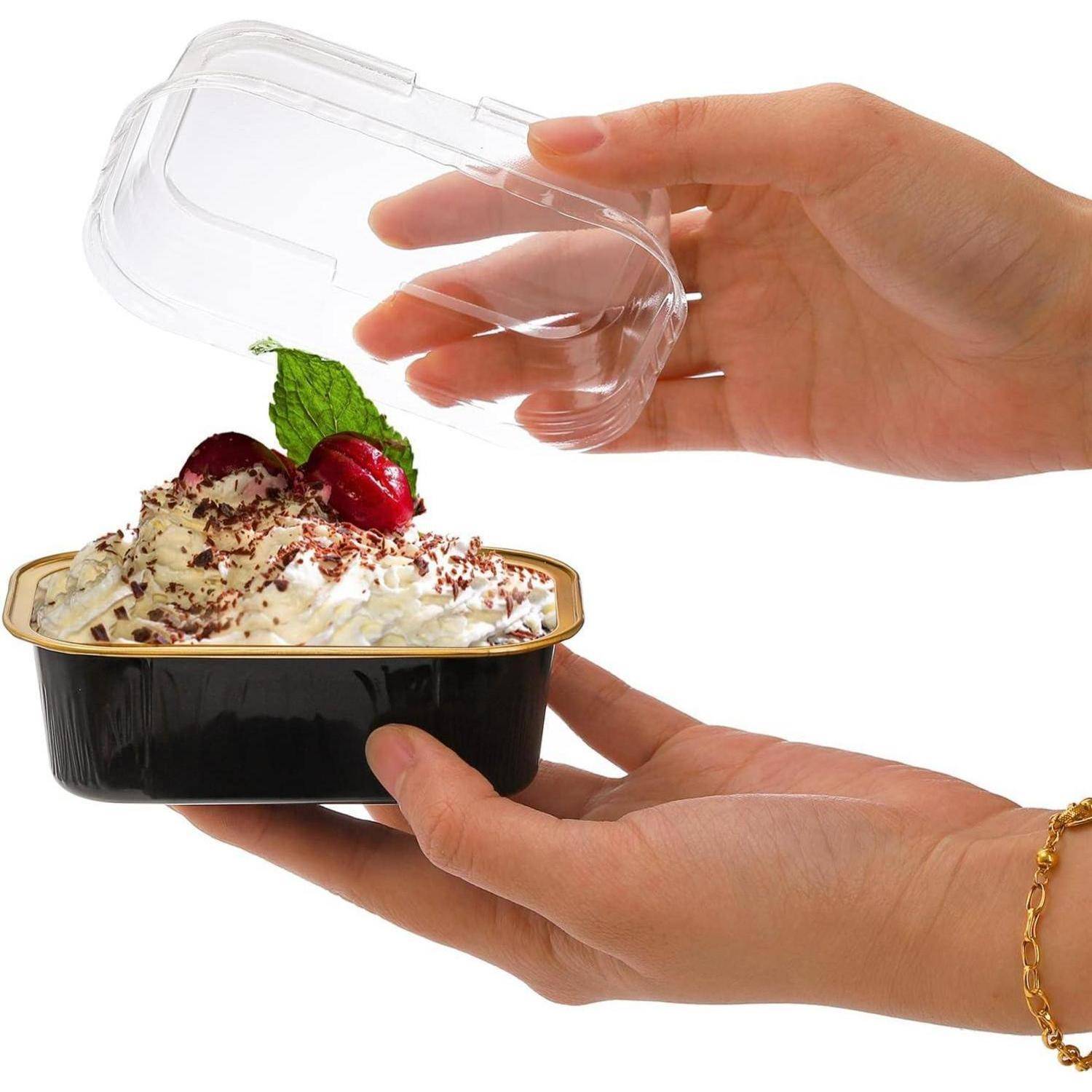 Eine Hand, die einen Behälter mit Dessert öffnet, das mit Schlagsahne, Schokolade, Kirschen und Minzblättern garniert ist.