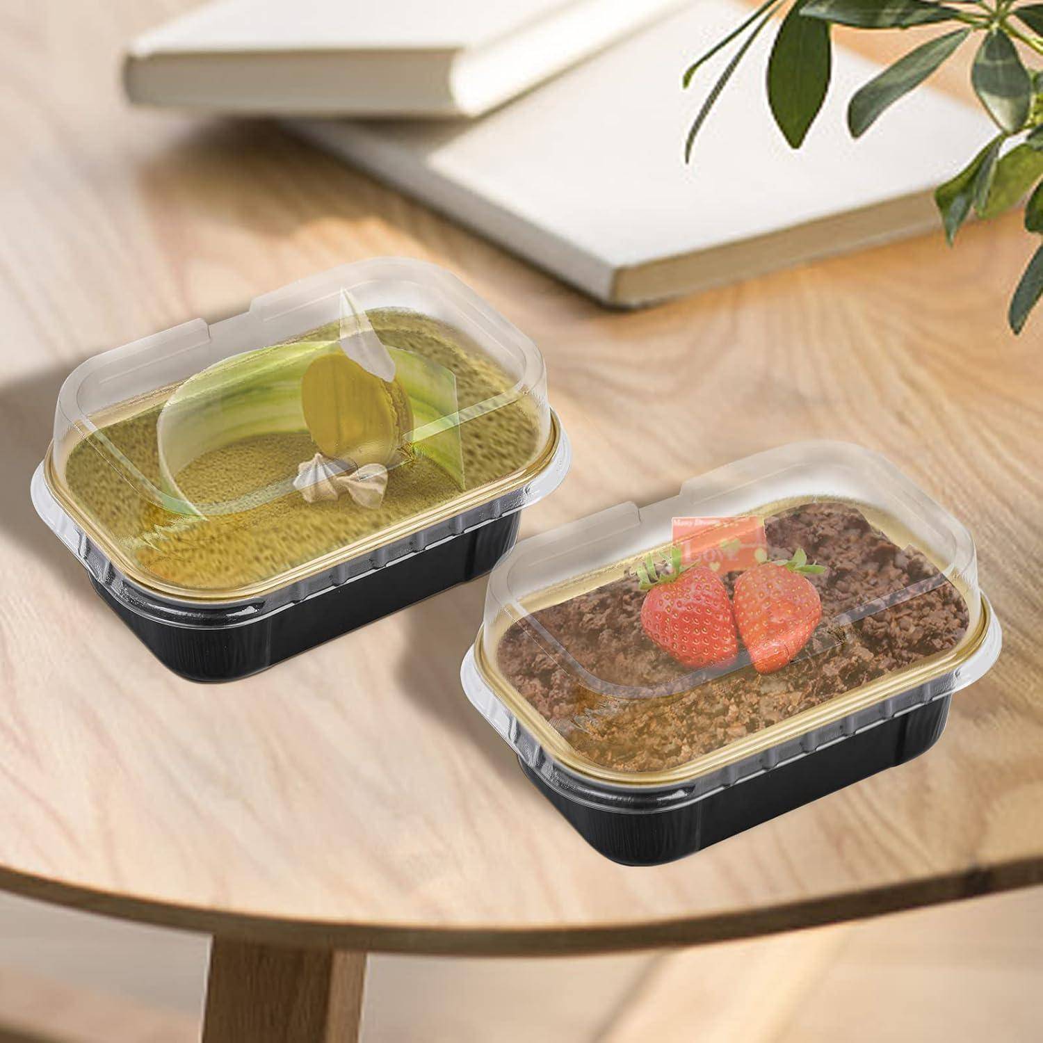 Zwei durchsichtige Plastikbehälter auf einem Holztisch, einer mit einer grünen Flüssigkeit und einem kleinen Schmuckstück, der andere mit einem braunen Dessert, das mit Erdbeeren garniert ist.