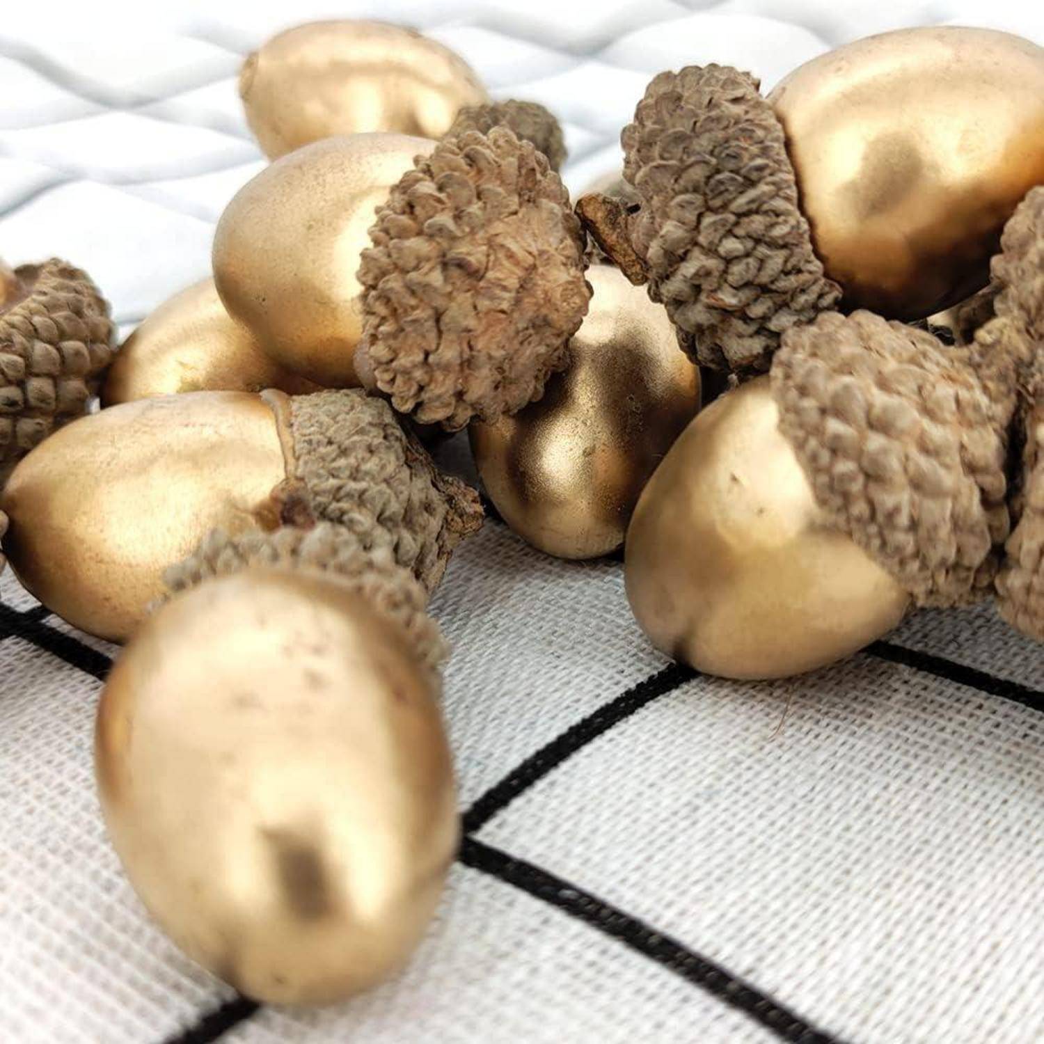 Goldene Eicheln mit strukturierten Käppchen sind auf einer weiß gemusterten Gitterfläche angeordnet, reflektieren das Licht und zeigen ihren glänzenden Finish.