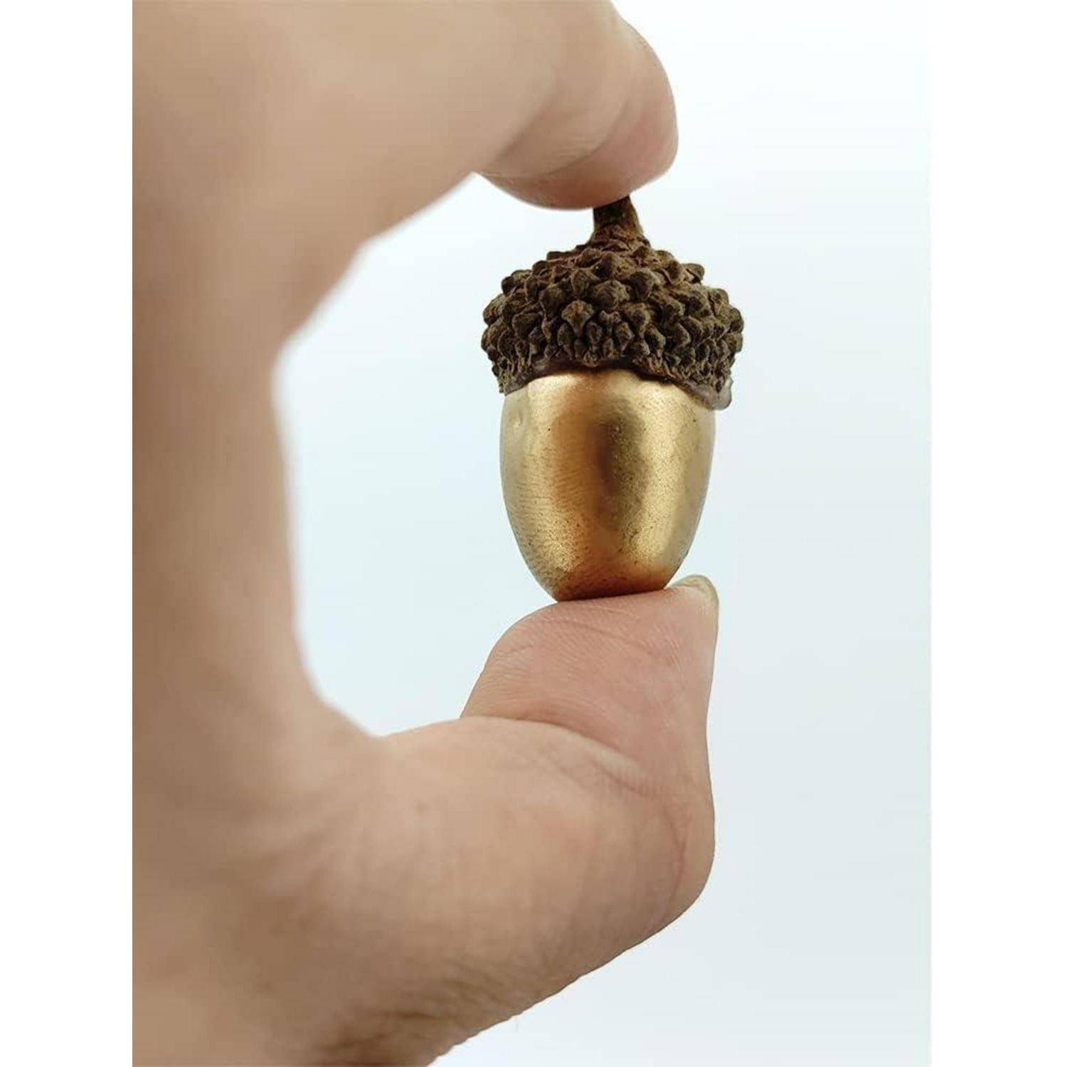 Eine Hand hält einen goldenen Eichel vor einem hellen Hintergrund, wobei der Kontrast zwischen der glänzenden Oberfläche der Eichel und ihrer strukturierten Kappe hervorgehoben wird.