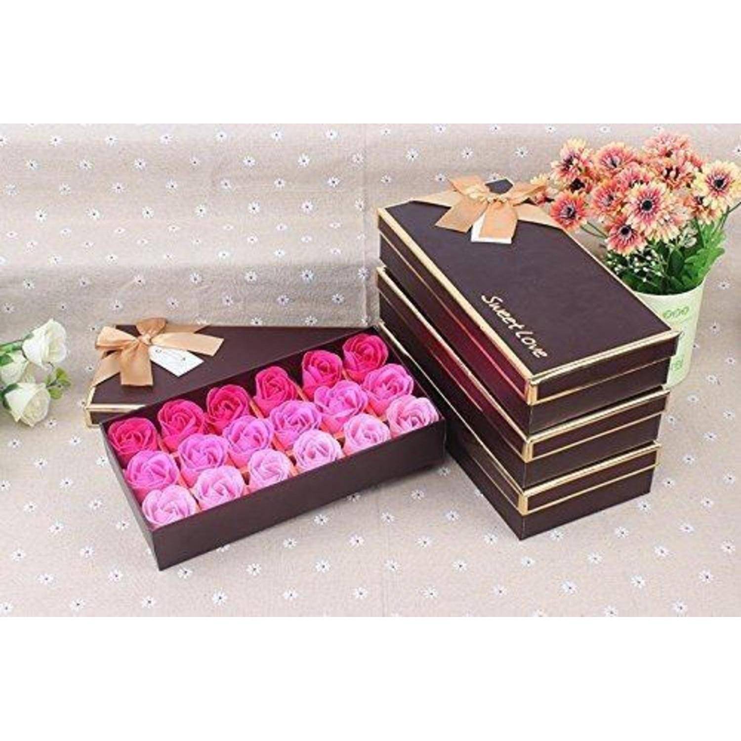 Geschenkboxen, gefüllt mit rosa und roten rosenförmigen Seifen, ordentlich angeordnet. Eine Box ist geöffnet, und die anderen sind daneben gestapelt, mit Blumen im Hintergrund.