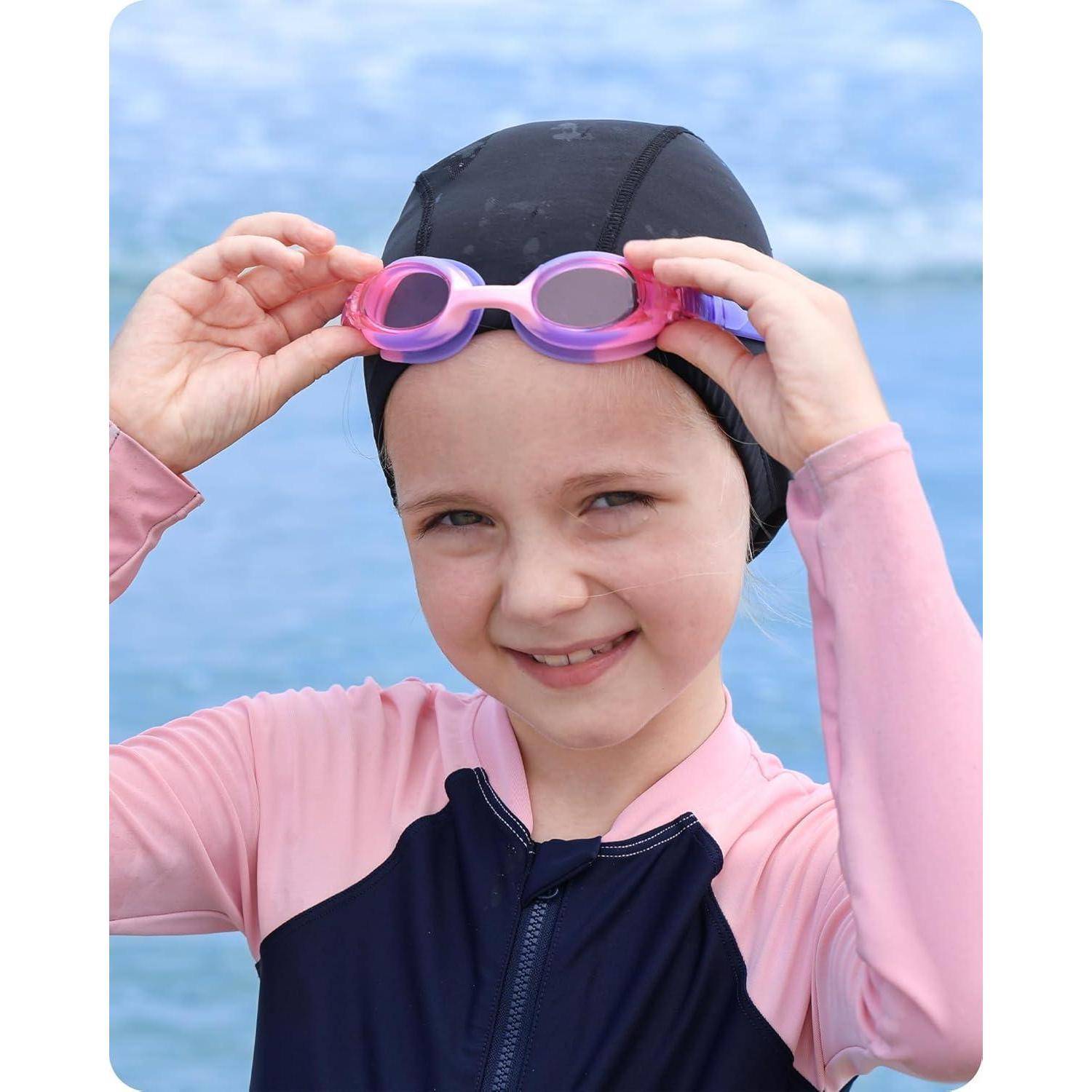 Ein junges Mädchen am Strand, das eine Badekappe trägt und ihre pinken Schwimmbrillen justiert, lächelt in die Kamera mit dem Ozean im Hintergrund.