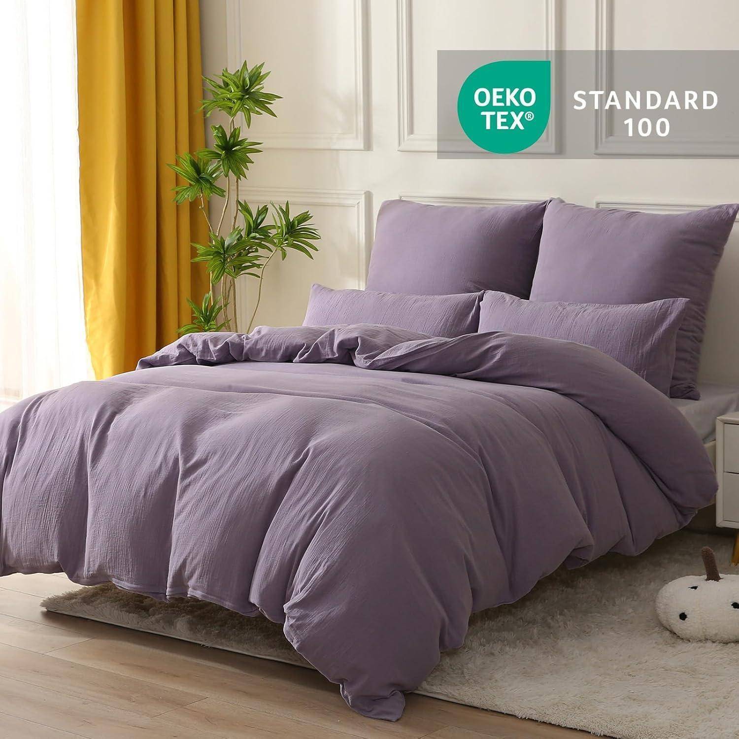 Ein sorgfältig gemachtes Bett mit Lavendelfarben Bettwäsche und Kissen steht in einem hellen Raum. Eine grüne Pflanze befindet sich neben dem Bett. Das 'OEKO-TEX STANDARD 100' Etikett ist sichtbar.
