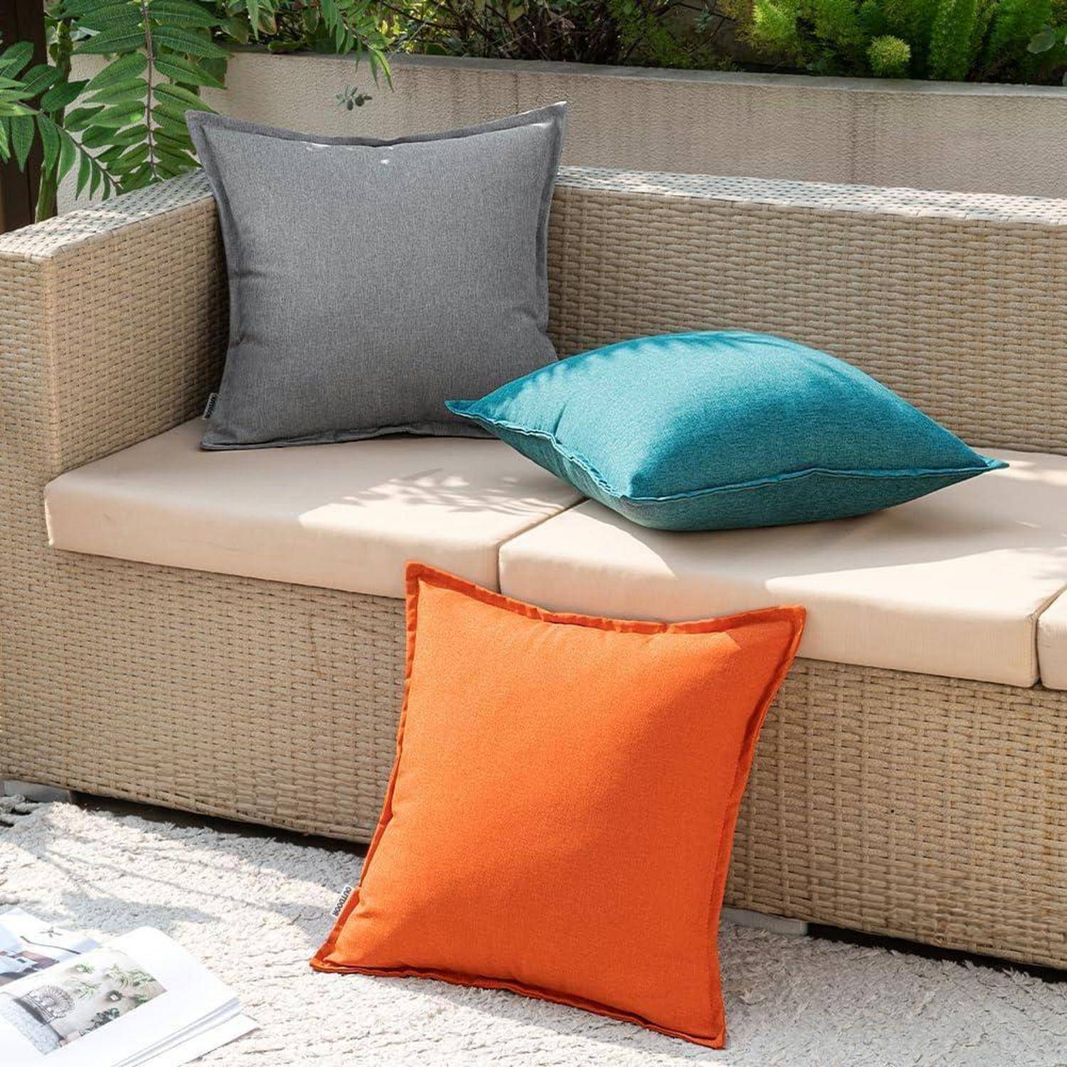 Ein beiges Outdoor-Sofa mit grauen, petrolblauen und orangefarbenen Kissen auf einem strukturierten Teppich, umgeben von Grünpflanzen.