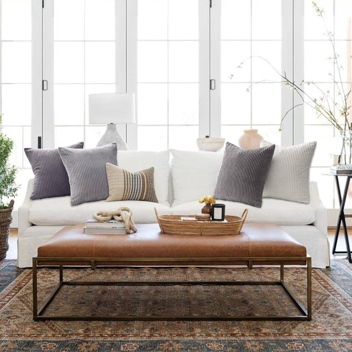 Ein moderner Wohnraum mit einem weißen Sofa, verschiedenen gestreiften Kissen, einem braunen Lederhocker, einem Blumenkorb und großen Fenstern im Hintergrund.