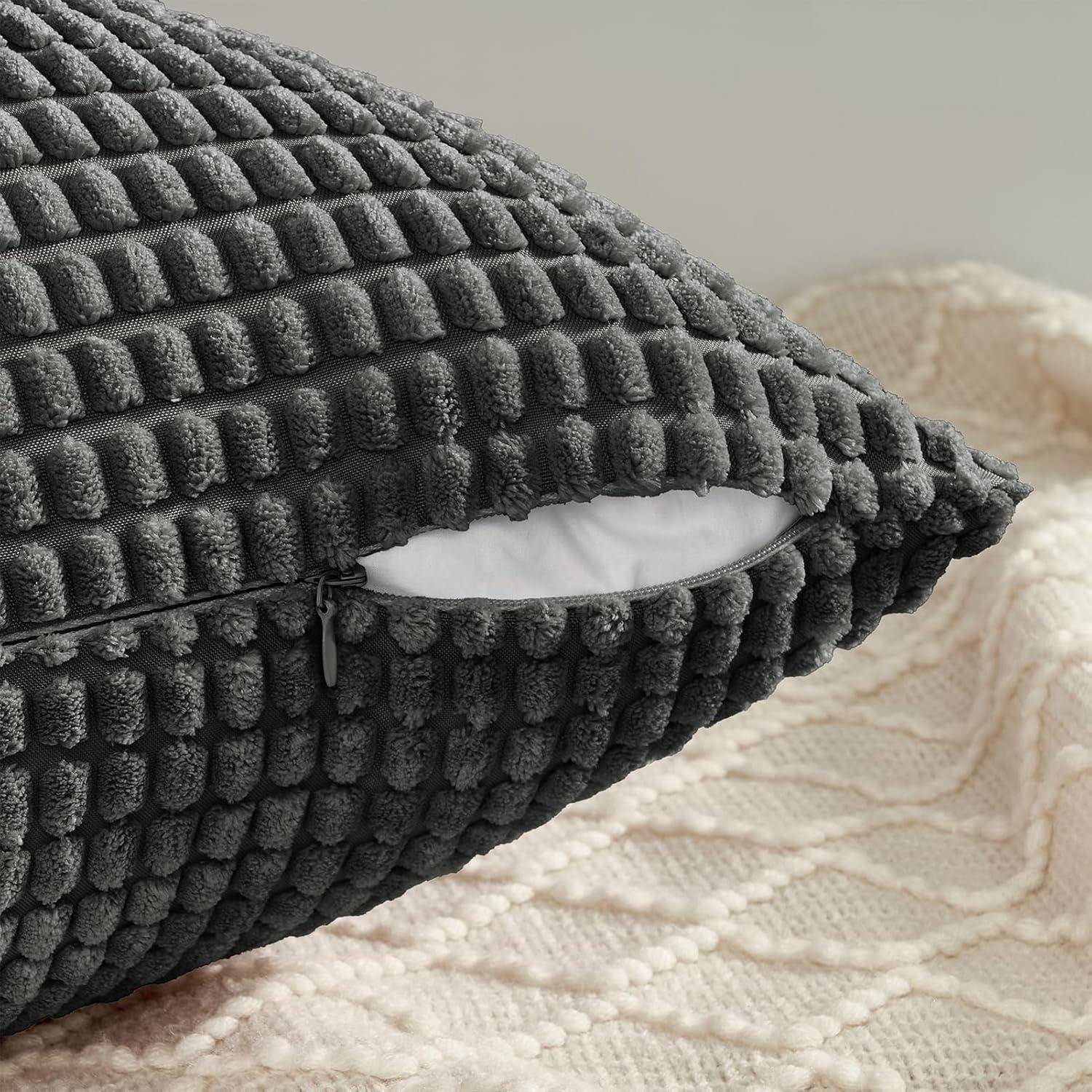 Dunkelgrauer strukturierter Kissenbezug mit sichtbarem Reißverschluss und weißer Füllung, auf einer cremefarbenen gestrickten Decke platziert.