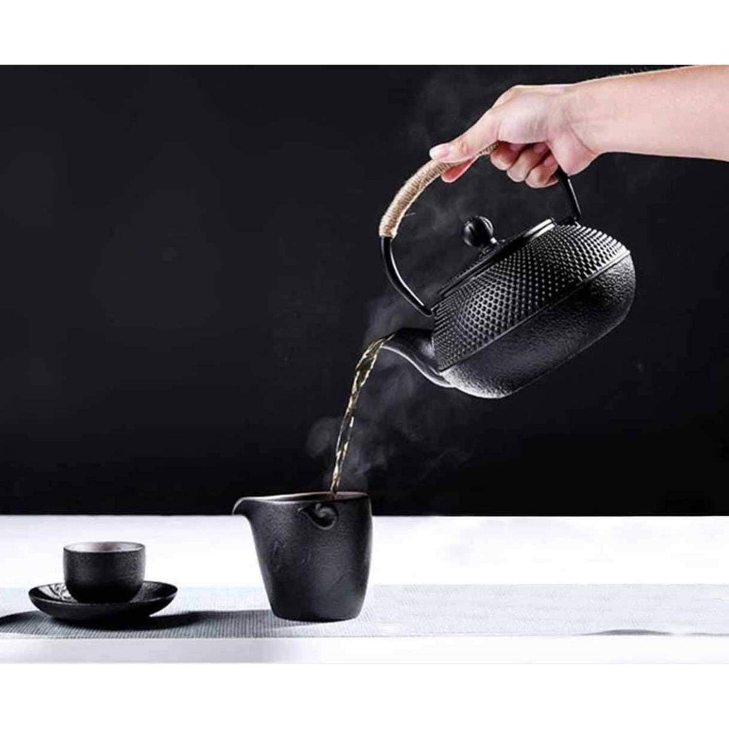 Eine Hand gießt Tee aus einer schwarzen Teekanne in eine Tasse, neben einer weiteren Tasse und Untertasse auf einem Tisch vor dunklem Hintergrund.
