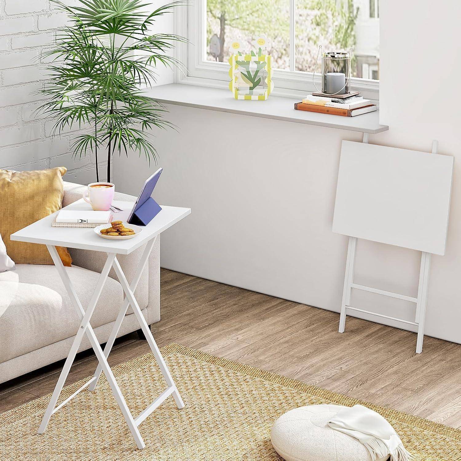 Ein gemütliches Wohnzimmer mit einem weißen zusammenklappbaren Tisch, auf dem ein Laptop, eine Tasse und ein Notizbuch stehen, neben einem Sofa mit Kissen. Eine Pflanze und eine leere Staffelei befinden sich in der Nähe.