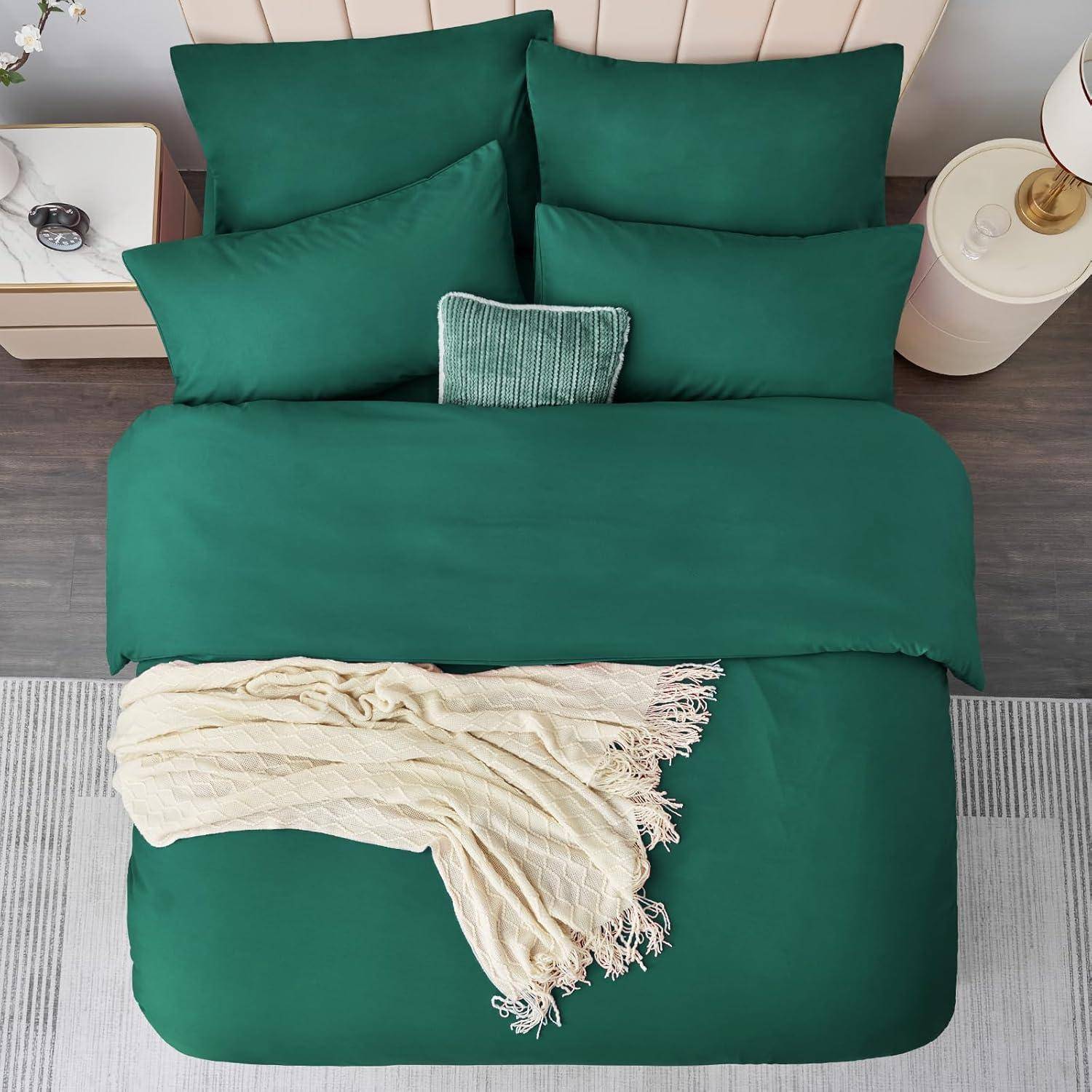 Ein ordentlich gemachtes Bett mit dunkelgrüner Bettwäsche, fünf Kissen und einer beigen Decke auf einem grauen Teppich. Ein Nachttisch trägt eine Lampe und eine Topfpflanze.