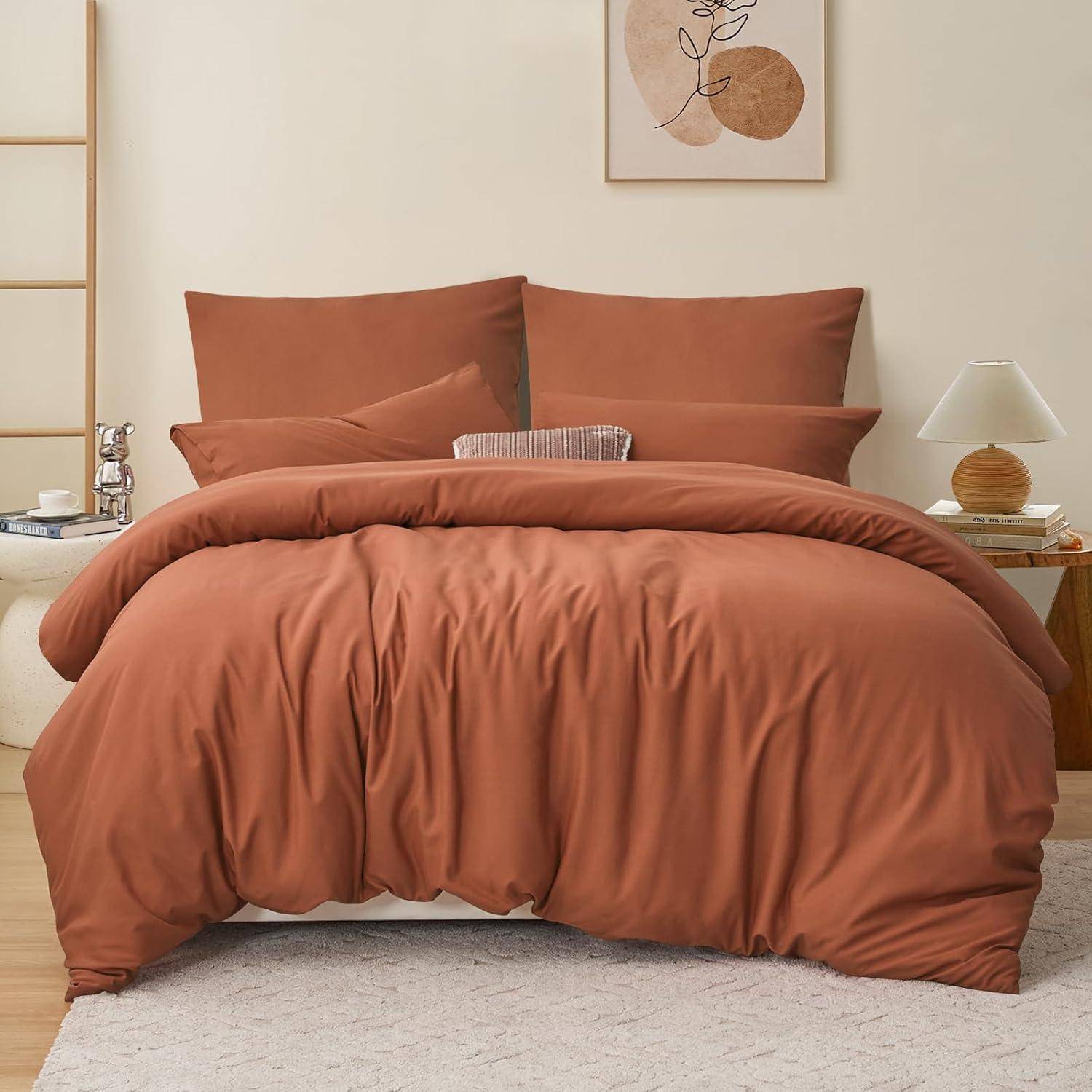 Ein ordentlich gemachtes Bett mit rostfarbener Bettwäsche und Kissen auf einem cremefarbenen Teppich in einem minimalistischen Schlafzimmer. Eine kleine Lampe und Deko stehen auf einem Beistelltisch.