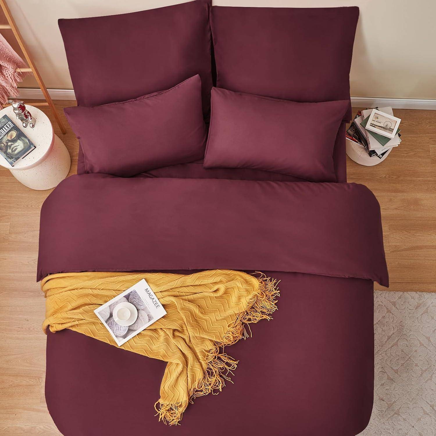 Ein ordentlich gemachtes Bett mit dunkelroten Laken und Kissen, akzentuiert durch eine gelbe Decke und Bücher auf dem Nachttisch, in einer gemütlichen Schlafzimmeratmosphäre.