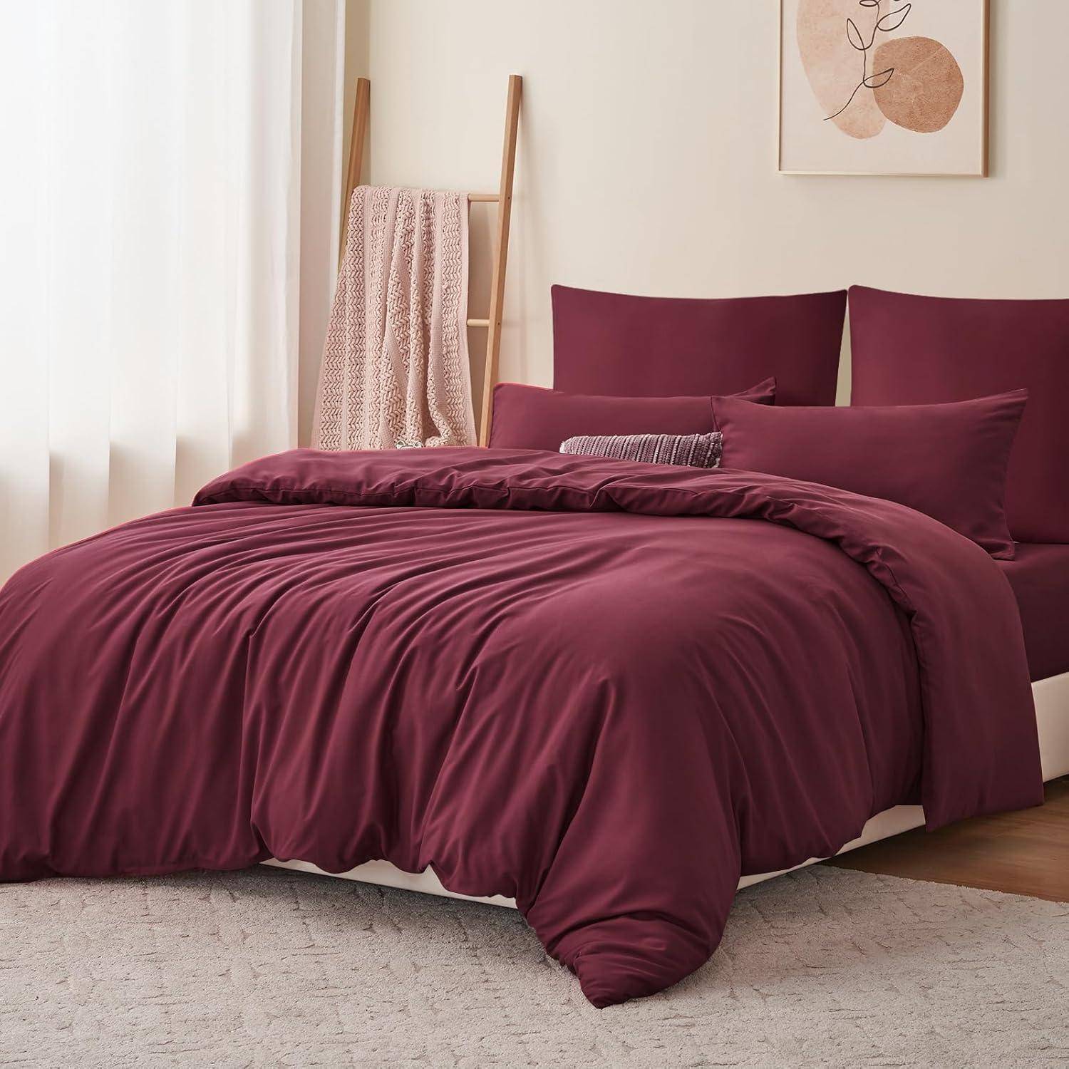 Ein Schlafzimmer zeigt ein ordentlich gemachtes Bett mit weinroter Bettwäsche und Kissen, eine Holzleiter mit einer beigen Decke und weiches, natürliches Licht.