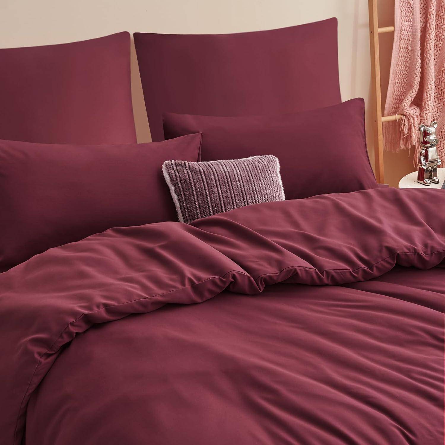 Ein kuscheliges Bett mit burgunderroten Laken und Kissen, versehen mit einem strukturierten grauen Kissen. Eine dekorative silberne Skulptur und eine rosa Decke sind sichtbar.