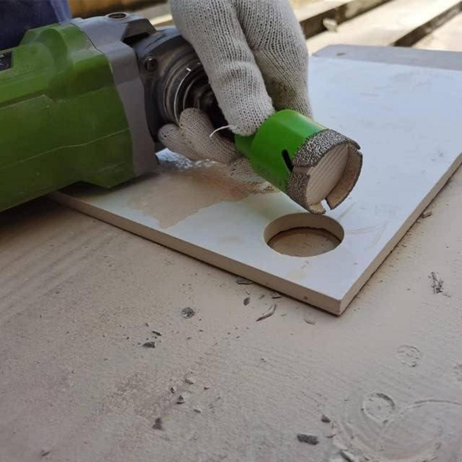 Eine Person mit Handschuhen verwendet eine grüne Bohrmaschine mit einer Lochsägeaufsatz, um ein kreisrundes Loch in eine flache, weiße Platte zu bohren.
