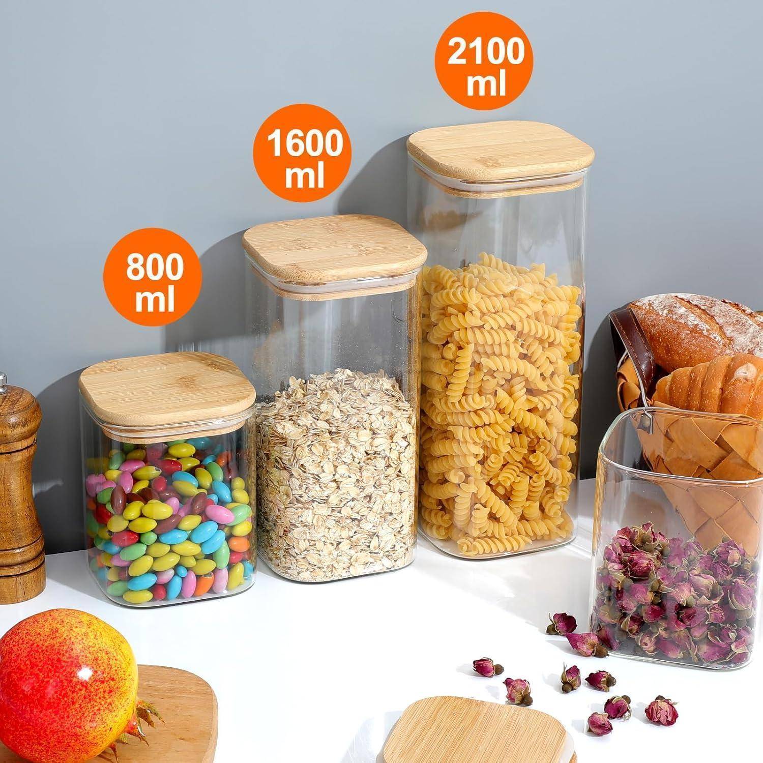 Vier transparente Behälter mit Bambus-Deckeln, beschriftet mit 800 ml, 1600 ml und 2100 ml, enthalten bunte Süßigkeiten, Haferflocken und Nudeln. Brot und Früchte in der Nähe.
