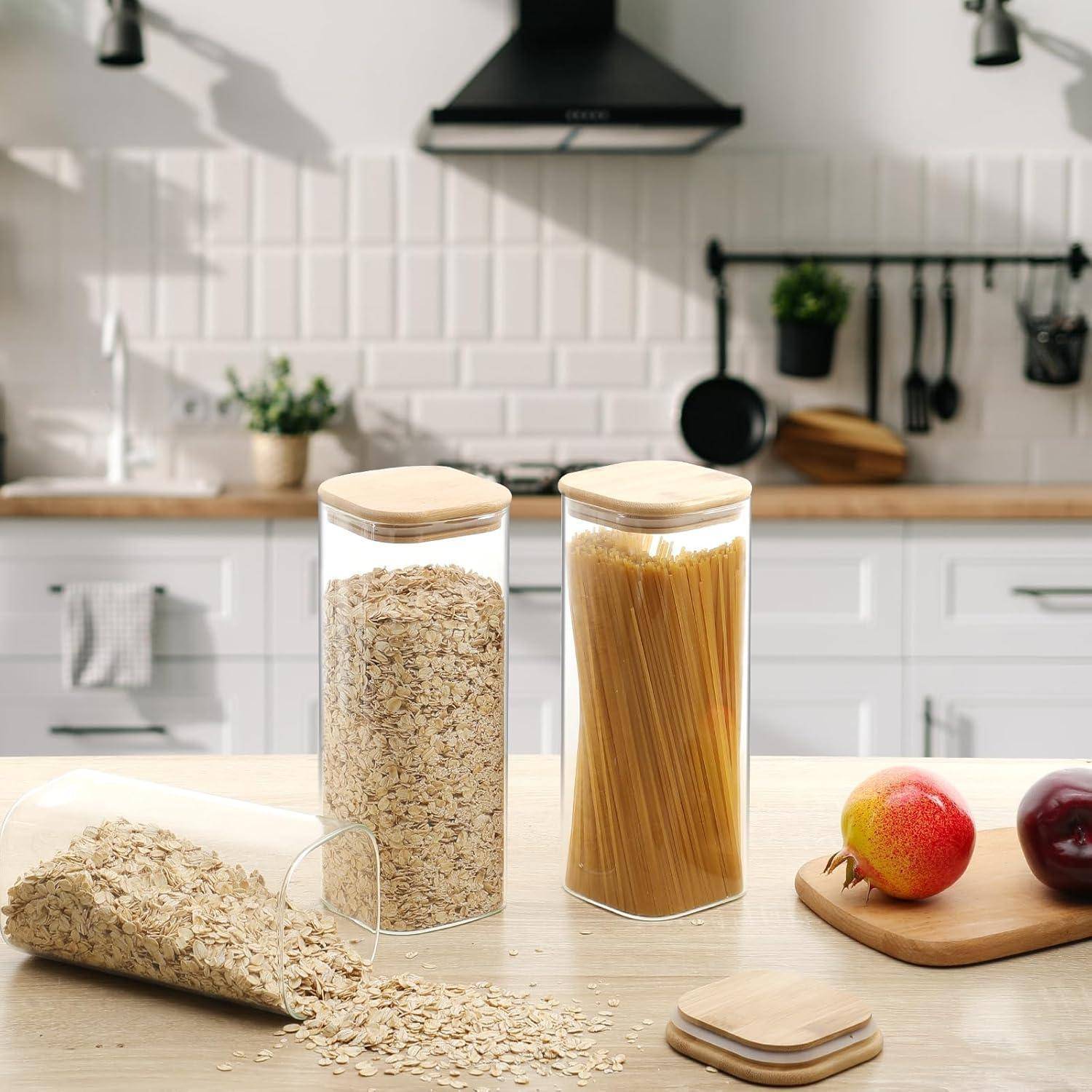 Küche mit Gläsern für Haferflocken und Spaghetti auf einer weißen Arbeitsplatte. Ein Glas ist umgekippt und lässt Haferflocken verschüttet. Frisches Obst und Küchenutensilien im Hintergrund.